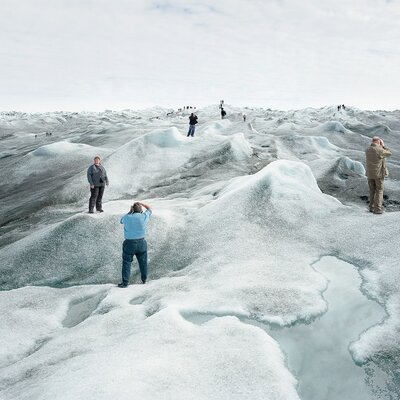 Eine Gruppe von Touristinnen und Touristen steht auf einer schmelzenden Eislandschaft und posiert an verschiedenen Punkten des Bildes für Fotos. An vielen Stellen der Eislandschaft hat sich schwarzer Ruß abgesetzt, das Eis ist dort sehr dunkel. Im Vordergrund ist eine große Pfütze aus Schmelzwasser.