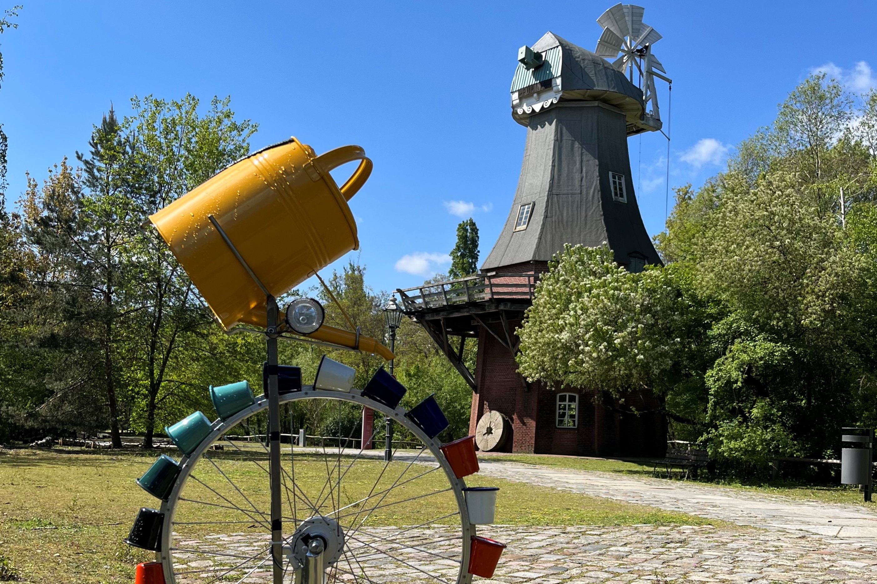 Ein Rad mit vielen bunten Bechern steht vor der Bockwindmühle im Museumspark. Über den Bechern ist eine Gieskanne befestigt.