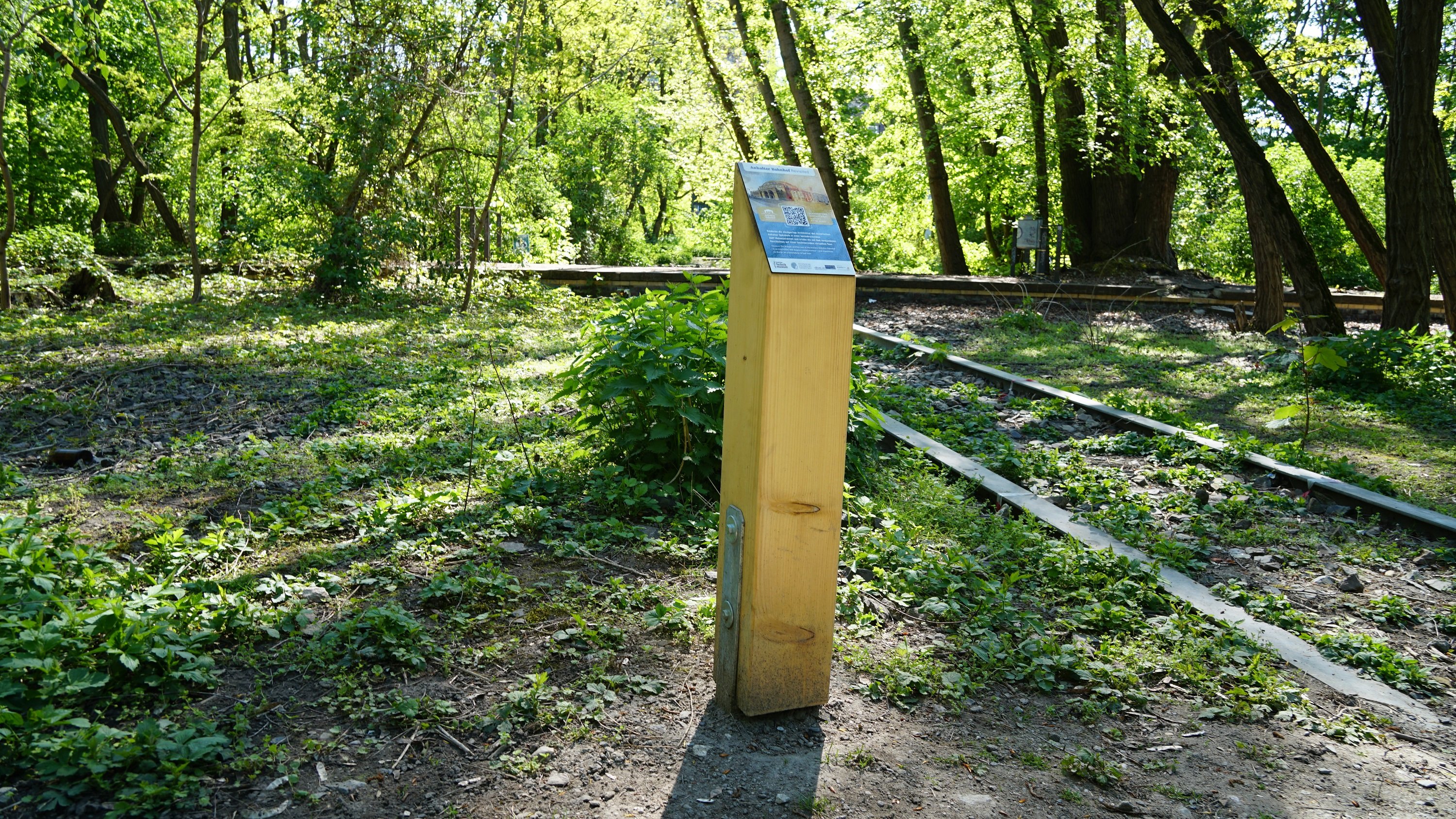 Eine Holstele in einem Park, darauf ein QR-Code. Es sind Schienen am Boden zu sehen.