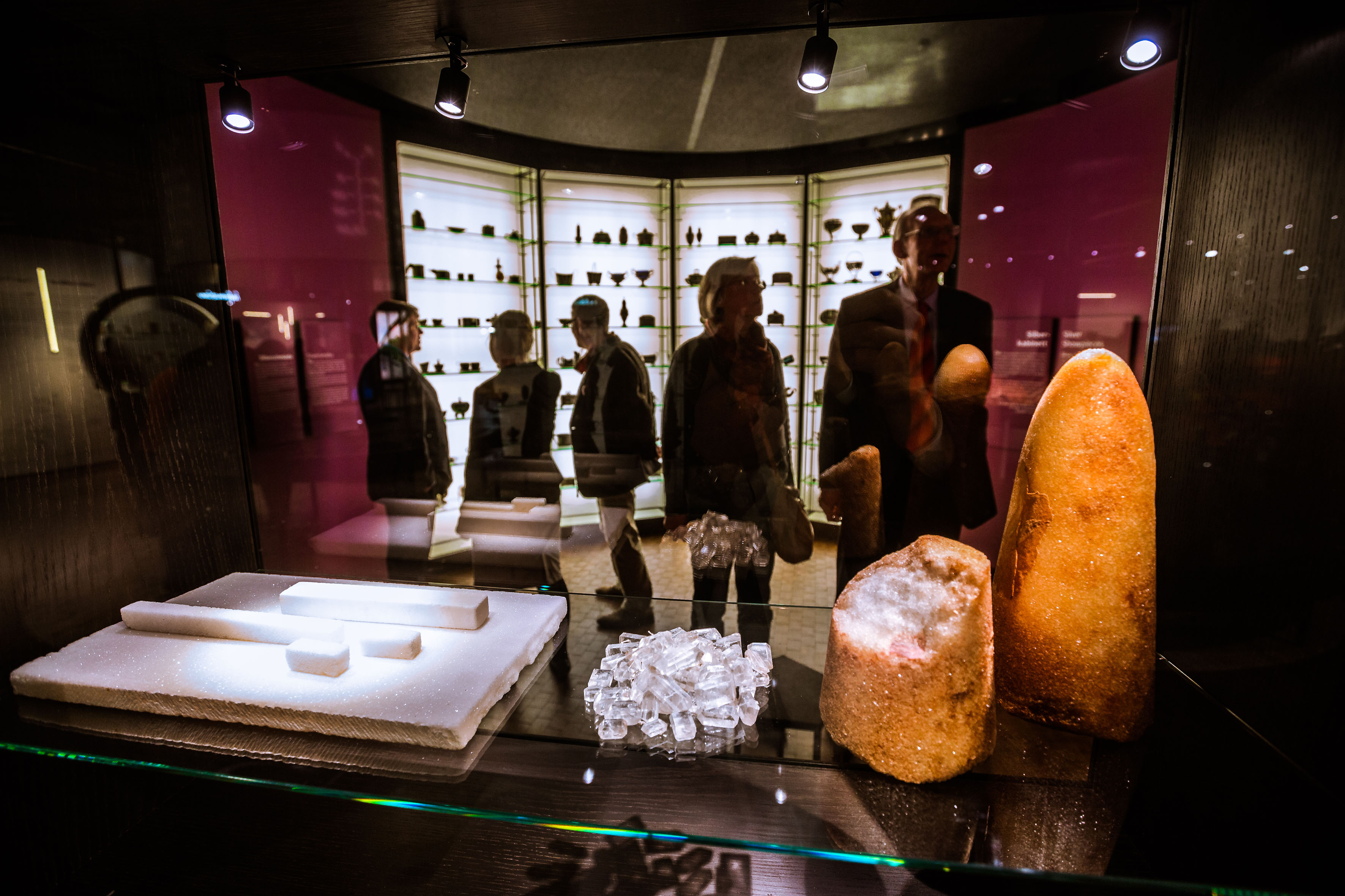 Fünf Personen stehen in einem Raum der Ausstellung „Alles Zucker!“. Im Vordergrund werden in einer Vitrine ein weiß-brauner Zuckerhut, Teile von Rohrzucker, Kandis-Zuckerkristalle und gepresster Zucker gezeigt. Im Hintergrund befinden sich deckenhohe Vitrinen mit Zuckerdosen.