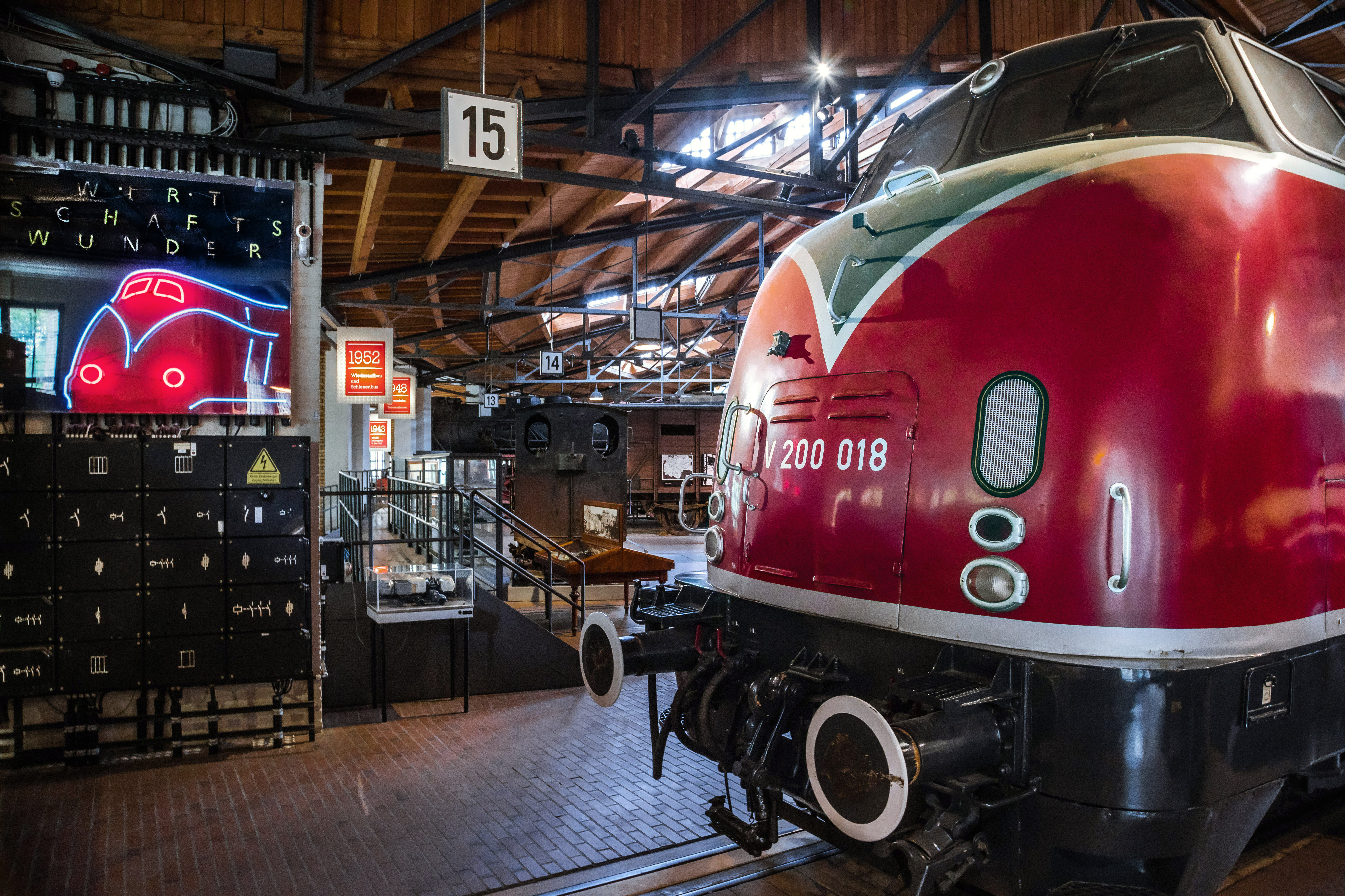 Die Schnellzuglokomotive V 200 steht in den historischen Lokschuppen des Deutschen Technikmuseums auf in den Holzboden eingelassenen Schienen. Ihr abgerundeter Rumpf ist Feuerrot lackiert. Links daneben befindet sich ein Neonschild in Form der Lokomotive. 