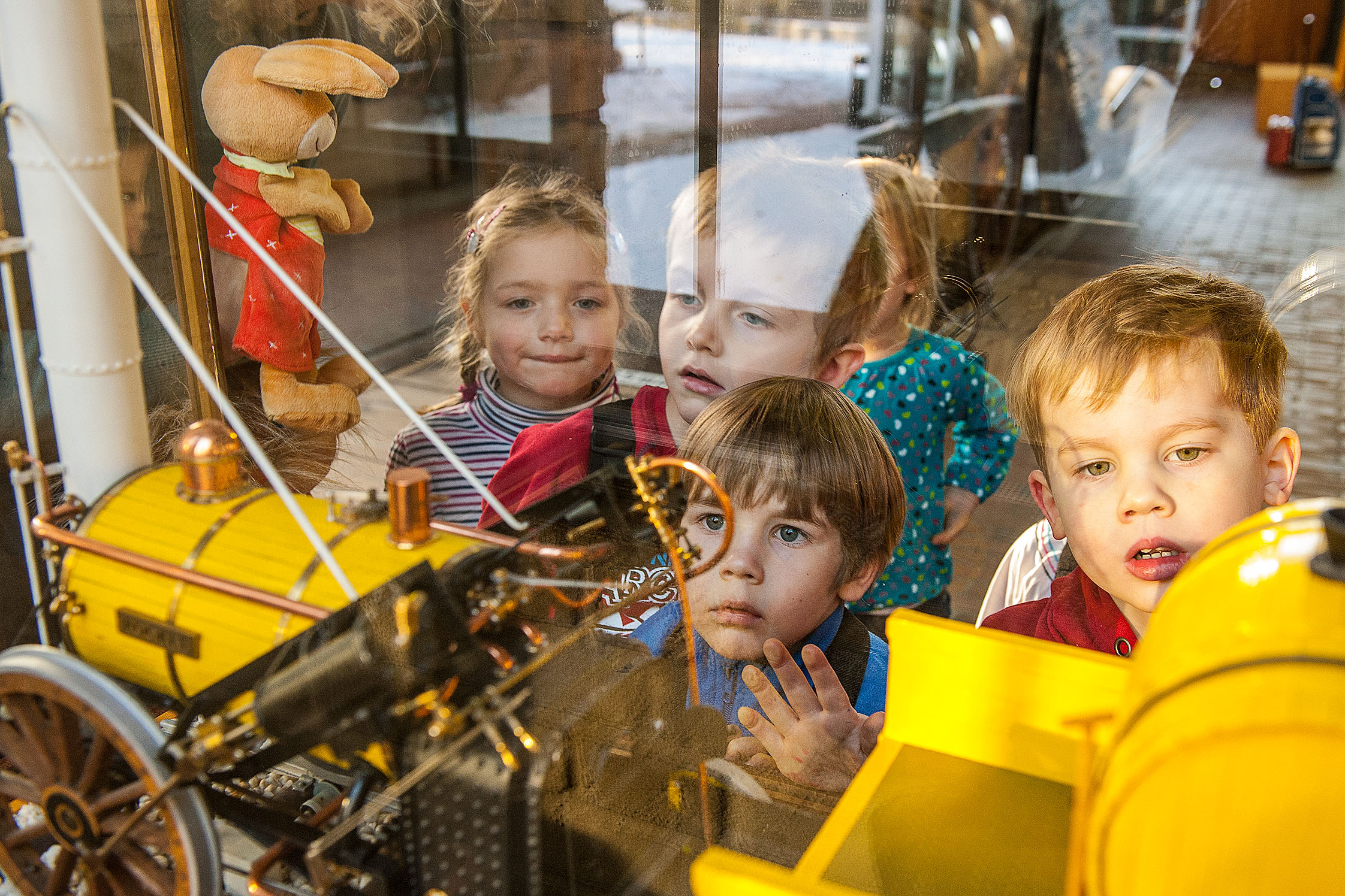 Fünf Kinder gucken in eine Vitrine, in der sich das Modell einer gelben Lokomotive befindet. Auf der linken Seite ist eine Hasen-Stoffpuppe zu sehen.