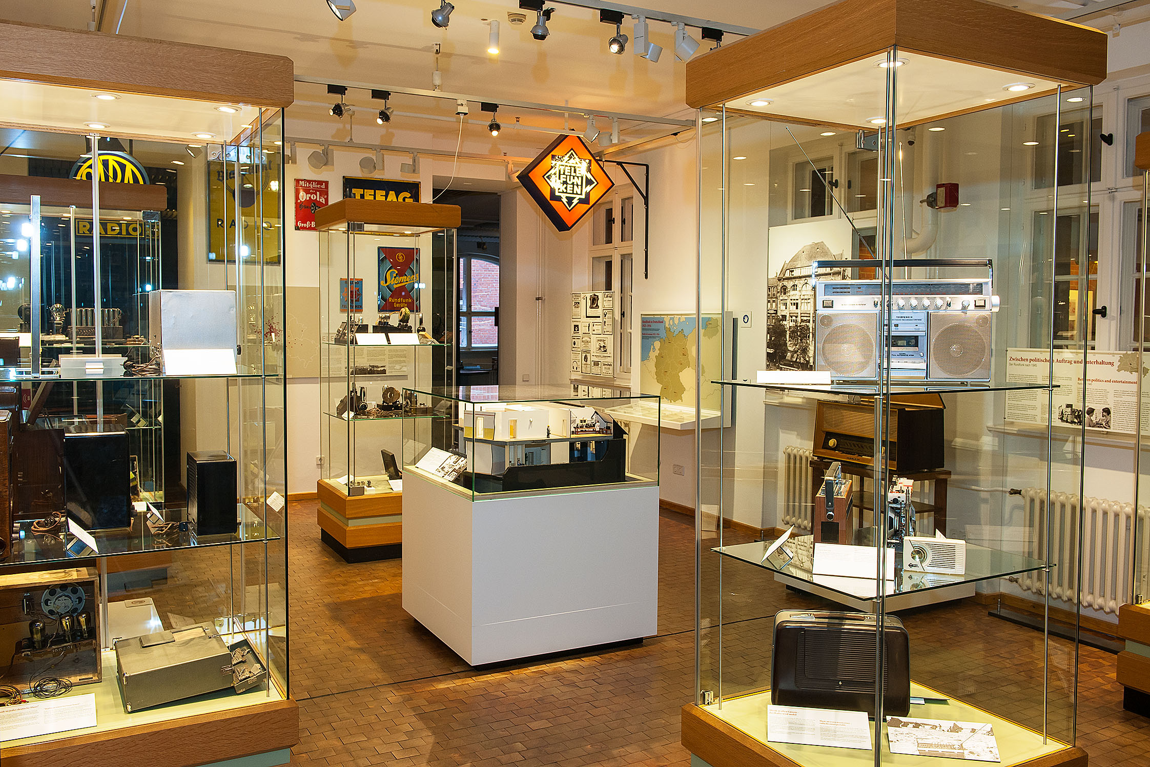 Mehrere Vitrinen in der Ausstellung zur Nachrichtentechnik. In den Vitrinen befinden sich auf mehreren Ebenen verschiedene Radiogeräte und Empfänger. Im Zentrum steht das Modell eines historischen Fernsehstudios, darüber hängt ein beleuchtetes Werbeschild.