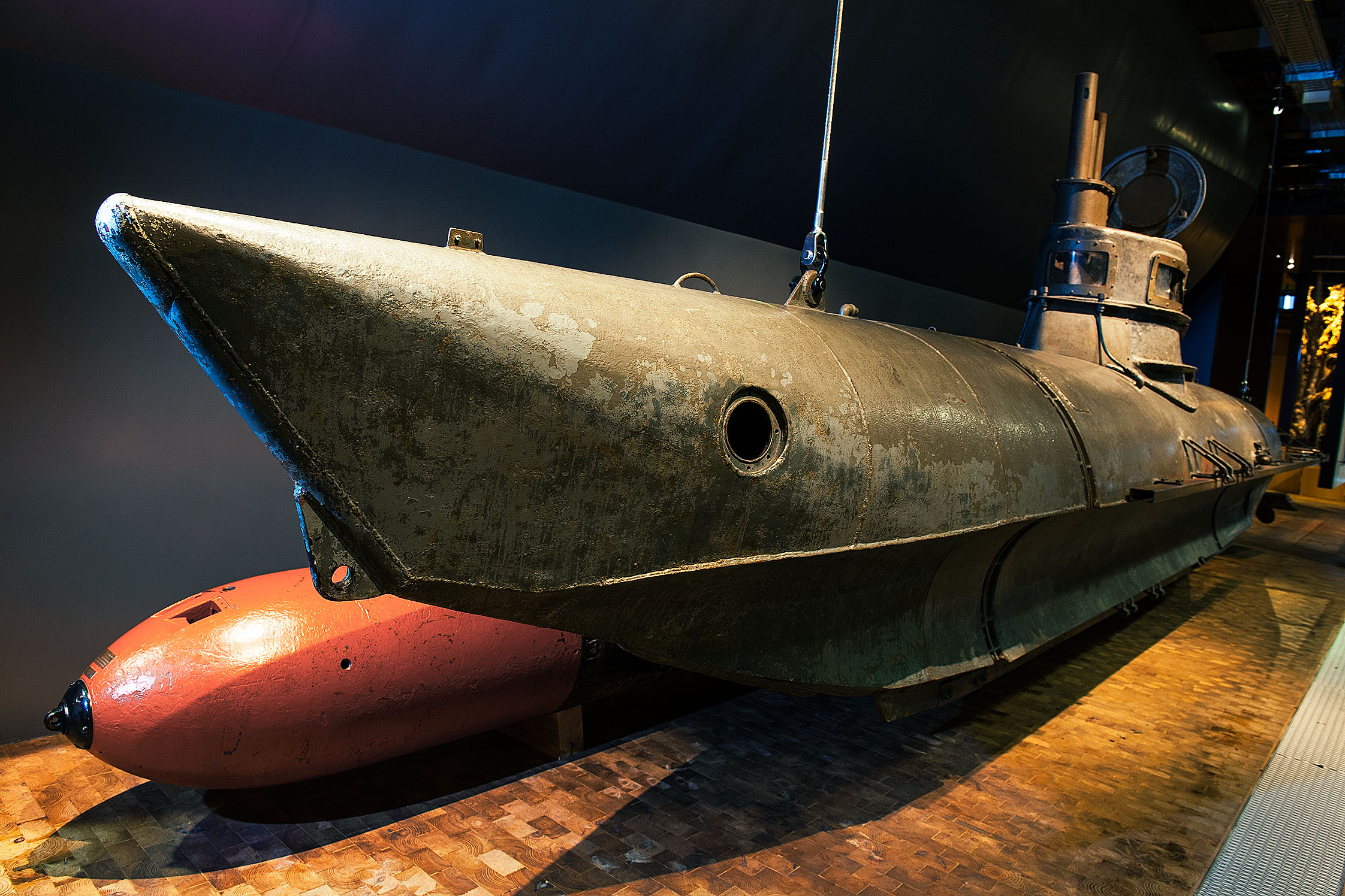 Schrägansicht des Ein-Mann-U-Boots Biber. Das U-Boot ist etwa neun Meter lang, verfügt über einen kleinen Turm mit Aussichtsfenstern und besteht aus grauem Metall. Die Nase des U-Boots ist nach oben gebogen. Dahinter liegt ein roter Torpedo.