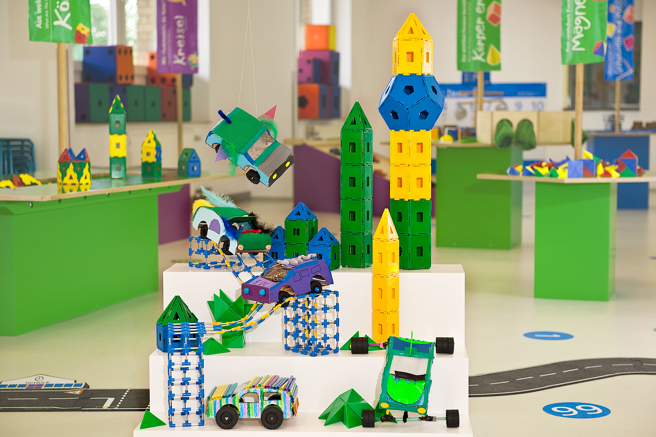 In einem Raum des Junior Campus stehen kleine selbstgebaute Fahrzeuge aus Pappe und Türme, die aus geometrischen Flächen aus Plastik zusammengesteckt wurden, auf einem dreistufigen Podest. Im Hintergrund befinden sich Arbeitstische mit weiteren Materialien.