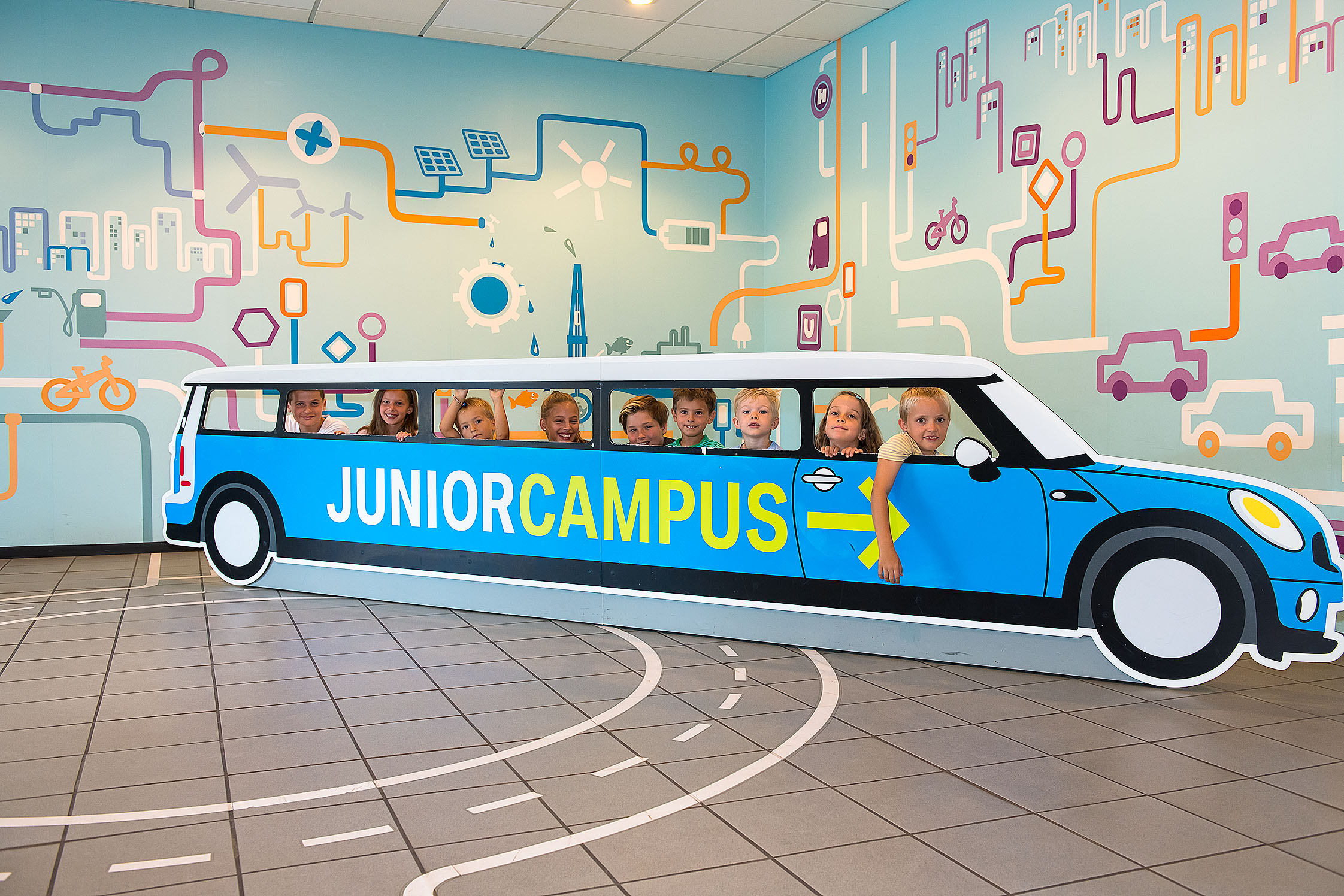 Neun Kinder schauen durch die Seitenfenster eines etwa sechs Meter langen Aufstellers aus Pappe in Form eines Autos. Auf der Seite ist der Schriftzug „Junior Campus“ in grün auf blauem Grund angebracht. Auf dem Boden sind Fahrbahnmarkierungen angedeutet.