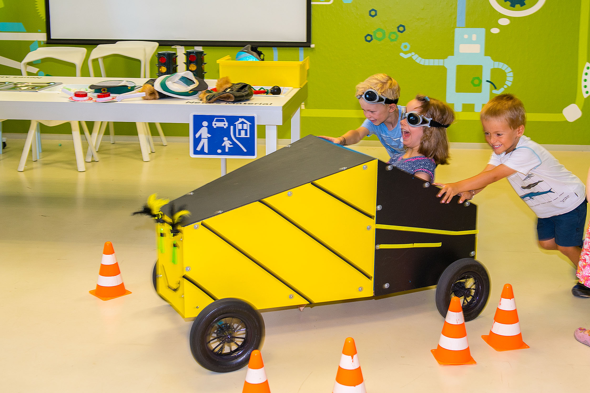 Drei Kinder fahren eine gelb-schwarze Seifenkiste. Das Fahrzeug besteht aus vorgefertigten Holzplatten mit vier Reifen. Auf die Front wurde ein lächelndes Gesicht gemalt. Ein Mädchen sitzt mit einer getönten Brille im Gefährt und steuert es, während zwei Jungen die Kiste anschieben.