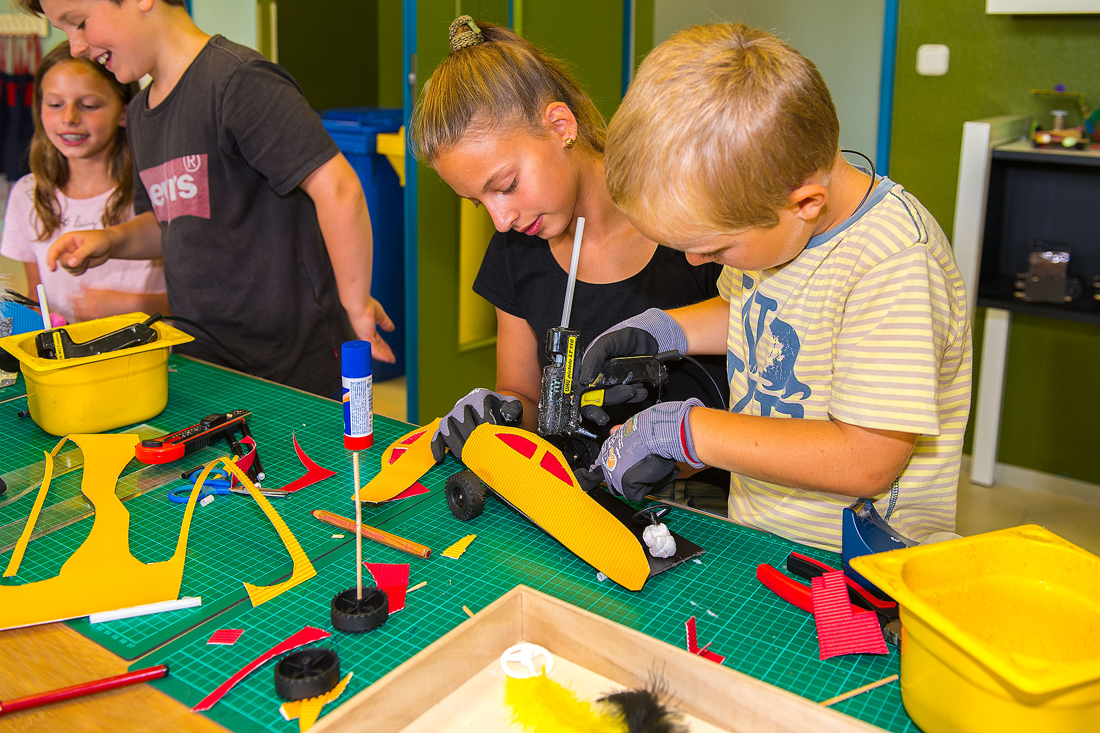 Vier Kinder kleben an einem Tisch kleine Autos aus Pappe mit einer Heißklebepistole zusammen. Auf dem Tisch liegen grüne Arbeitsmatten, bunte Pappschnipsel, gelbe Schalen und Klebestifte. 