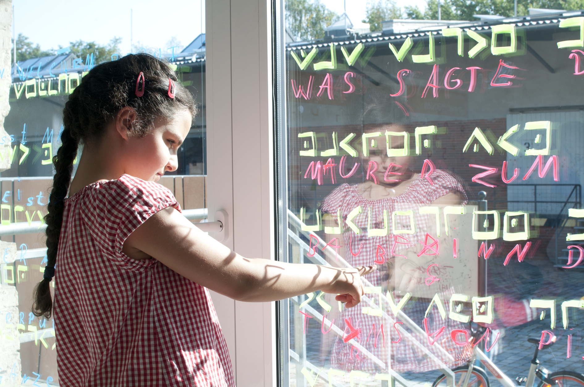 Ein Mädchen deutet auf eine Fensterscheibe, auf der in grün verschiedene Zeichen geschrieben sind. Darunter stehen in roter Farbe Buchstaben und Worte.