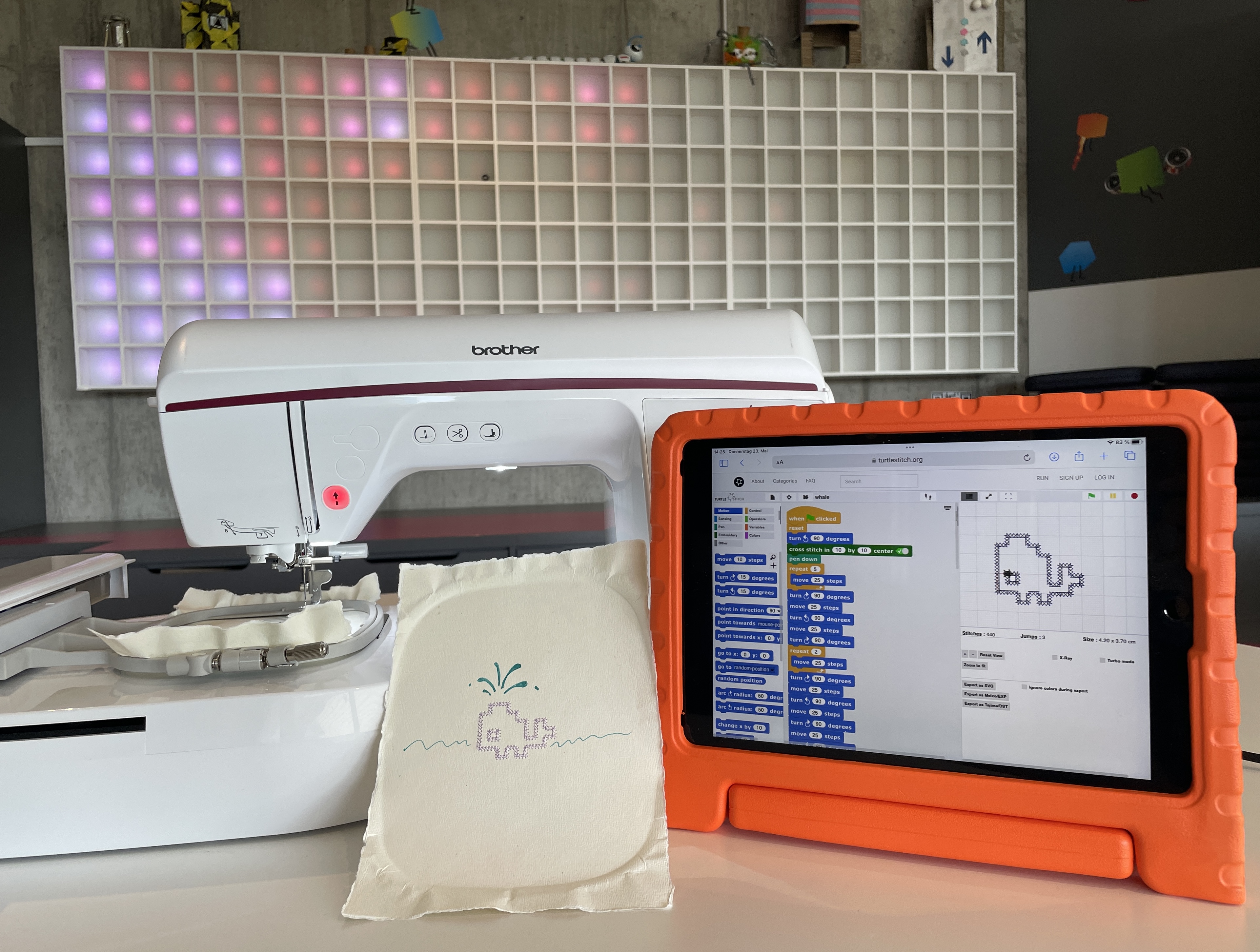 Eine Nahmaschine, ein gesticketes Bild von einem Wal und ein Bildschirm mit organgenem Rand auf dem eine Programmier-Software läuft.