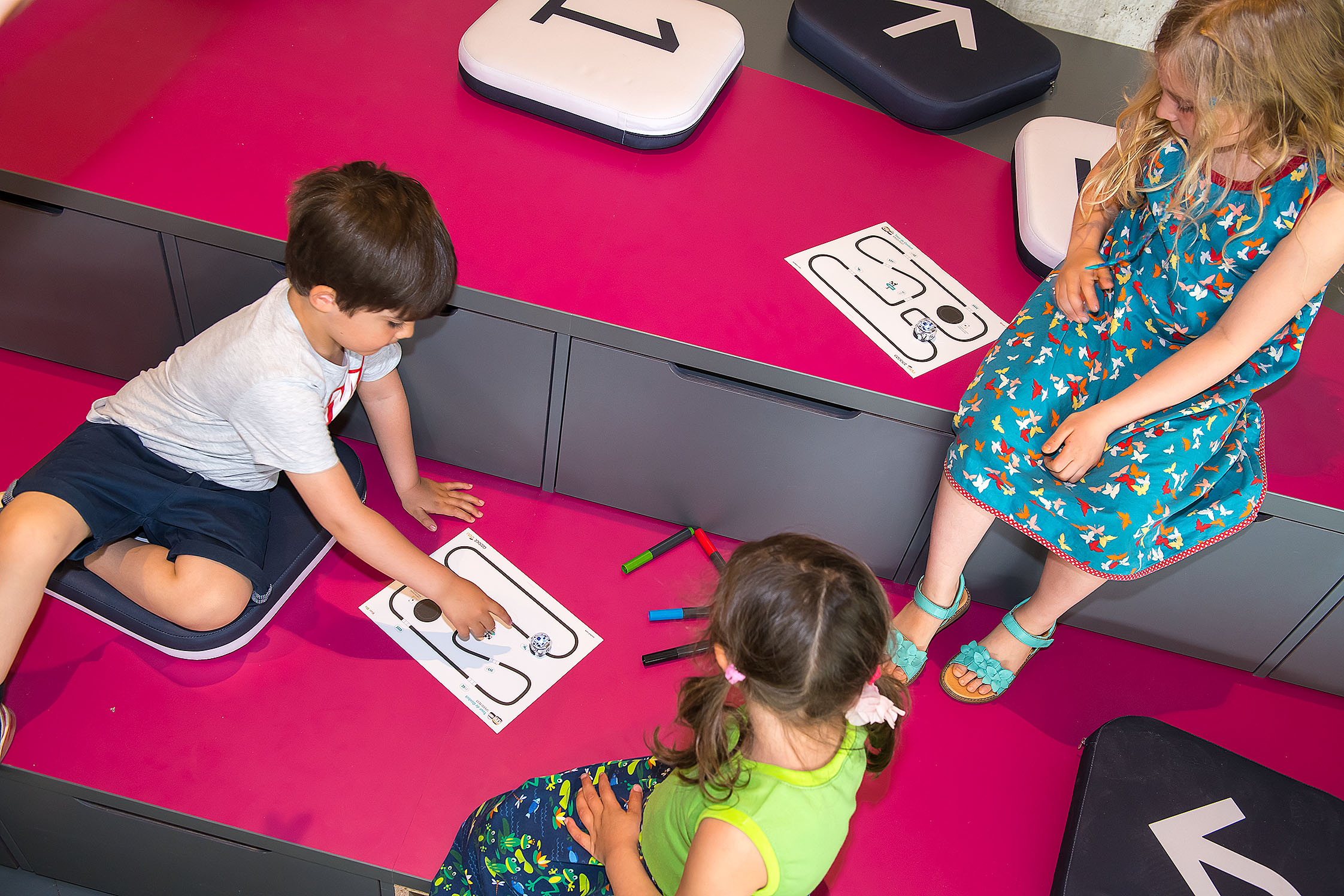 Ein kleiner Junge zeigt auf gemalte Linien, auf denen ein Robotik-Spielzeug entlangfährt, zwei Mädchen schauen zu. 