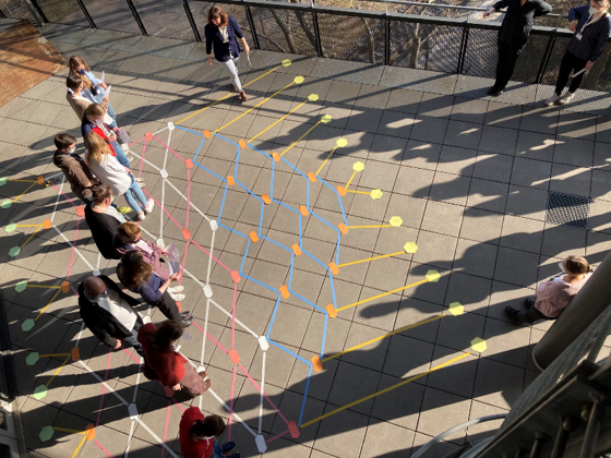 Eine Grupper erwachsener Personen steht auf einer Terrasse. Auf dem Boden ist ein Netz aus verschiedenen Farben aufgemalt.