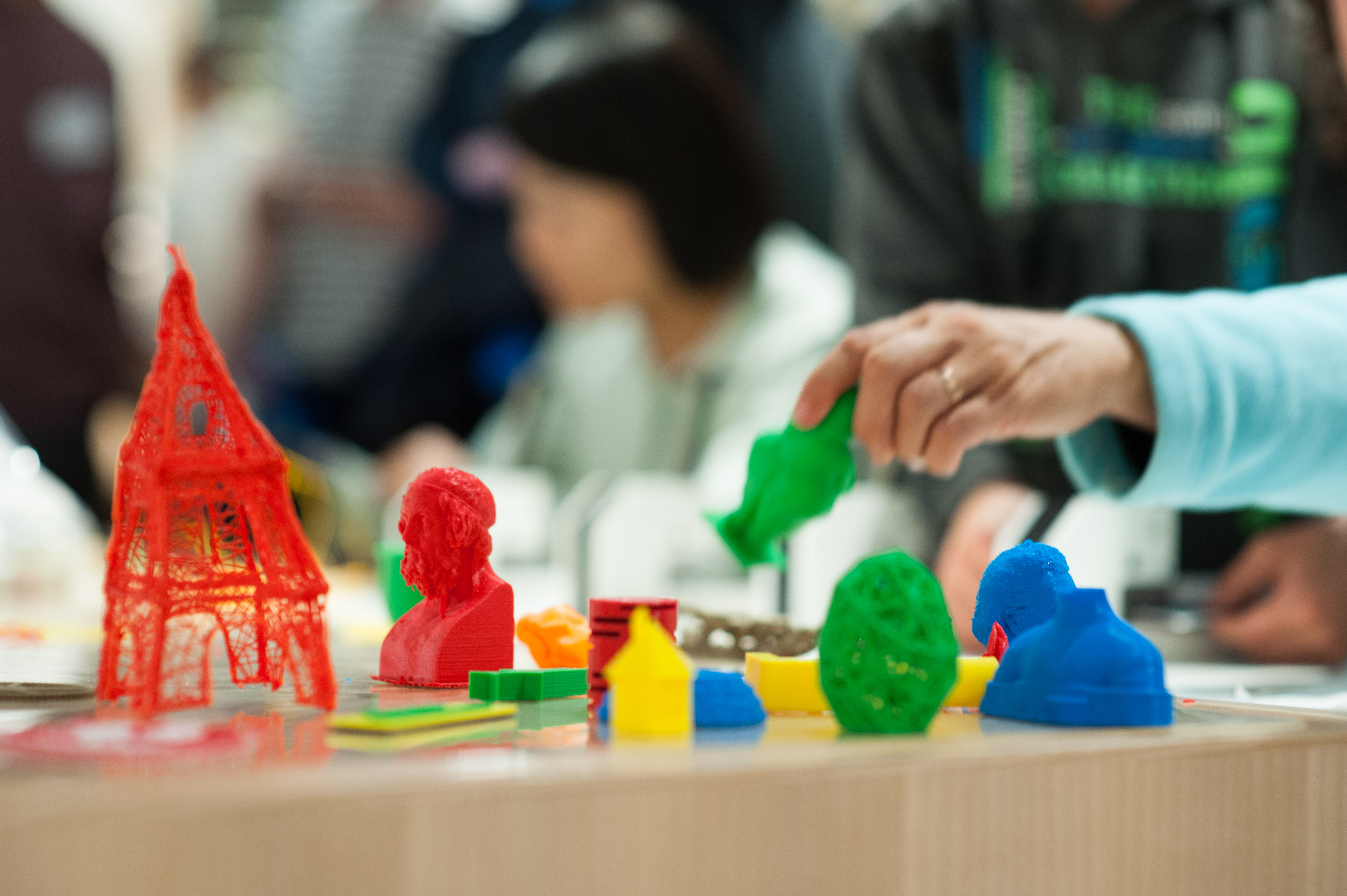 Auf einem Tisch stehen Figuren, Gegenstände und Kunstwerke, die mit einem 3D-Drucker aus verschiedenfarbigem, recyclebarem Plastik gedruckt wurden. Links steht eine rote, etwa 15 Zentimeter hohe, mit einem 3D-Stift gezeichnete, grobe Nachbildung des Eifelturms.