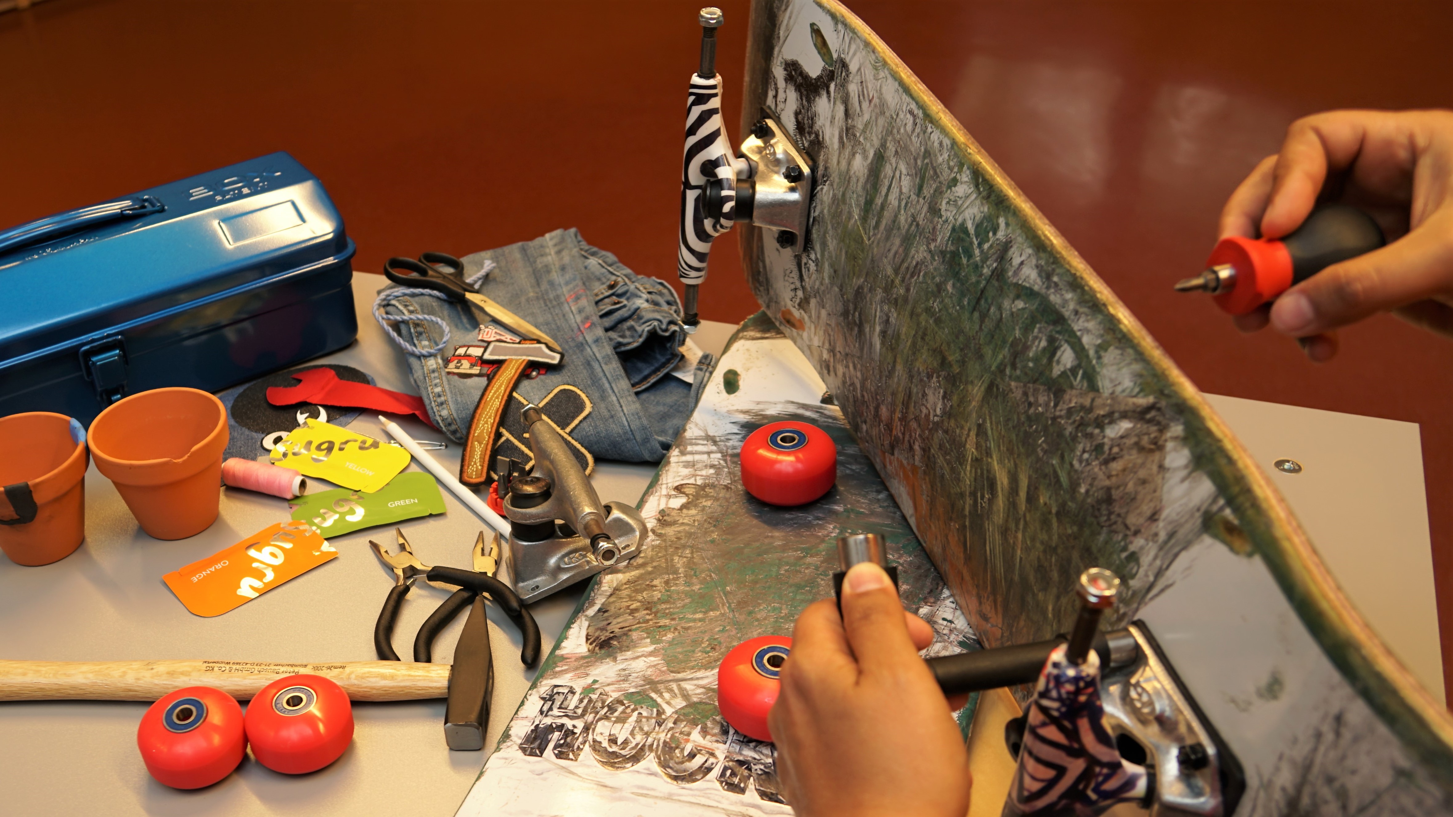 Auf einem Tisch liegen ein kaputtes Skateboard, kaputte Blumentöpfe und eine kaputte Jeans und die Hilfsmittel, um sie zu reparieren.