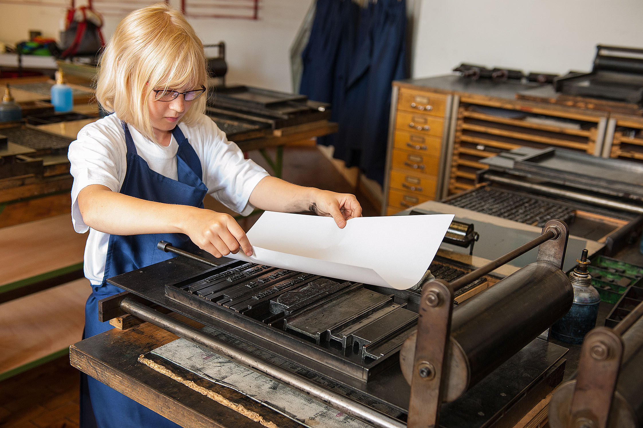 Ein Mädchen steht vor einer kleinen Druckerpresse und legt vorsichtig ein Blatt Papier auf einen gesetzten Text. Sie trägt eine Schürze und hält das Blatt behutsam mit einer Hand an jeder Seite fest.