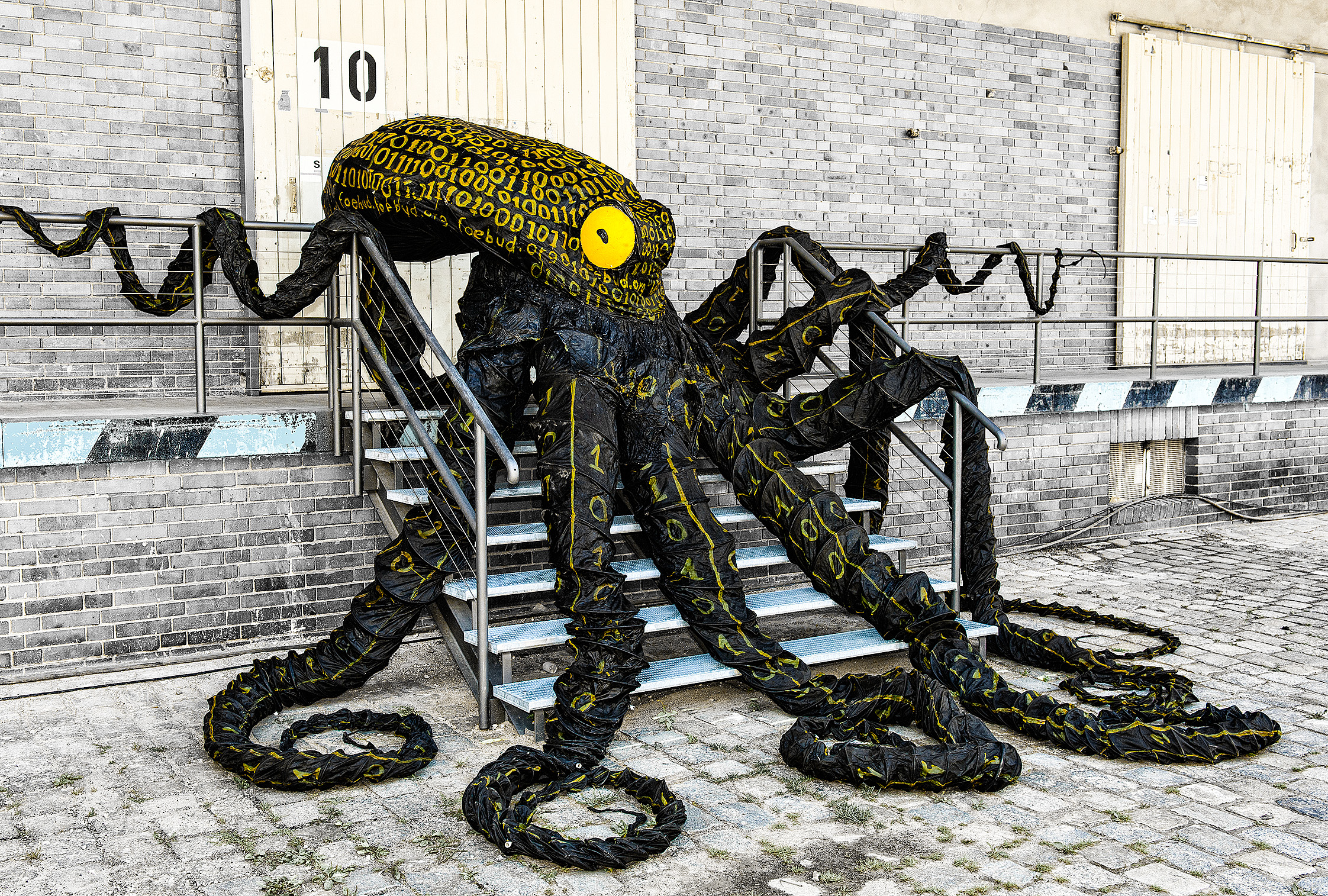 Der Datenkrake Otto liegt auf einer breiten Metalltreppe vor einem Holztor. Seine acht schwarzen Arme sind über die Stufen drapiert. Sein Auge leuchtet gelb. Auf Kopf und Armen sind viele gelbe Nullen und Einsen gemalt.