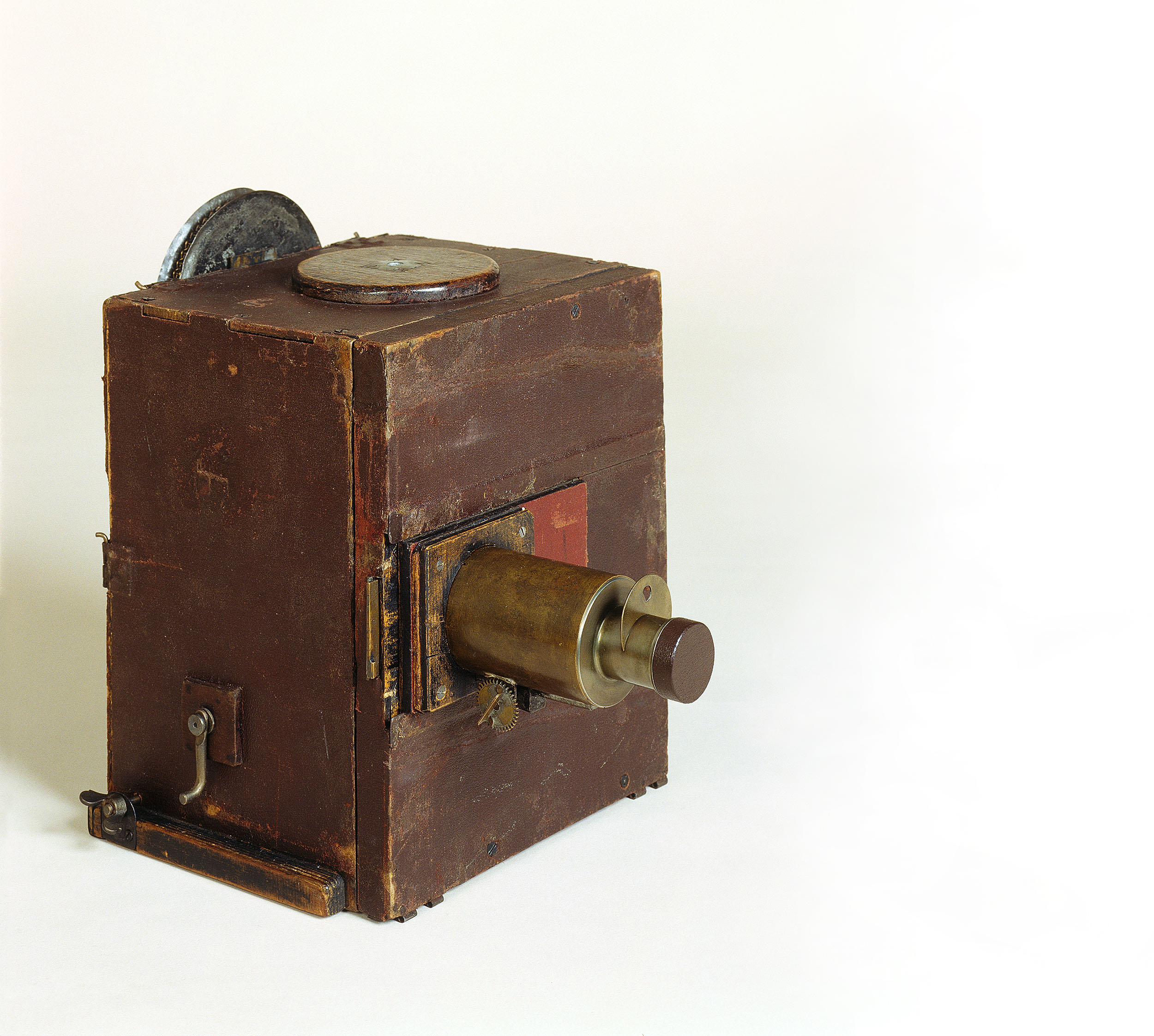 Die Kamera ist ein einfacher brauner Holzkasten. An der Vorderseite sitzt das Objektiv, an der rechten Seite befindet sich eine kleine Handkurbel.