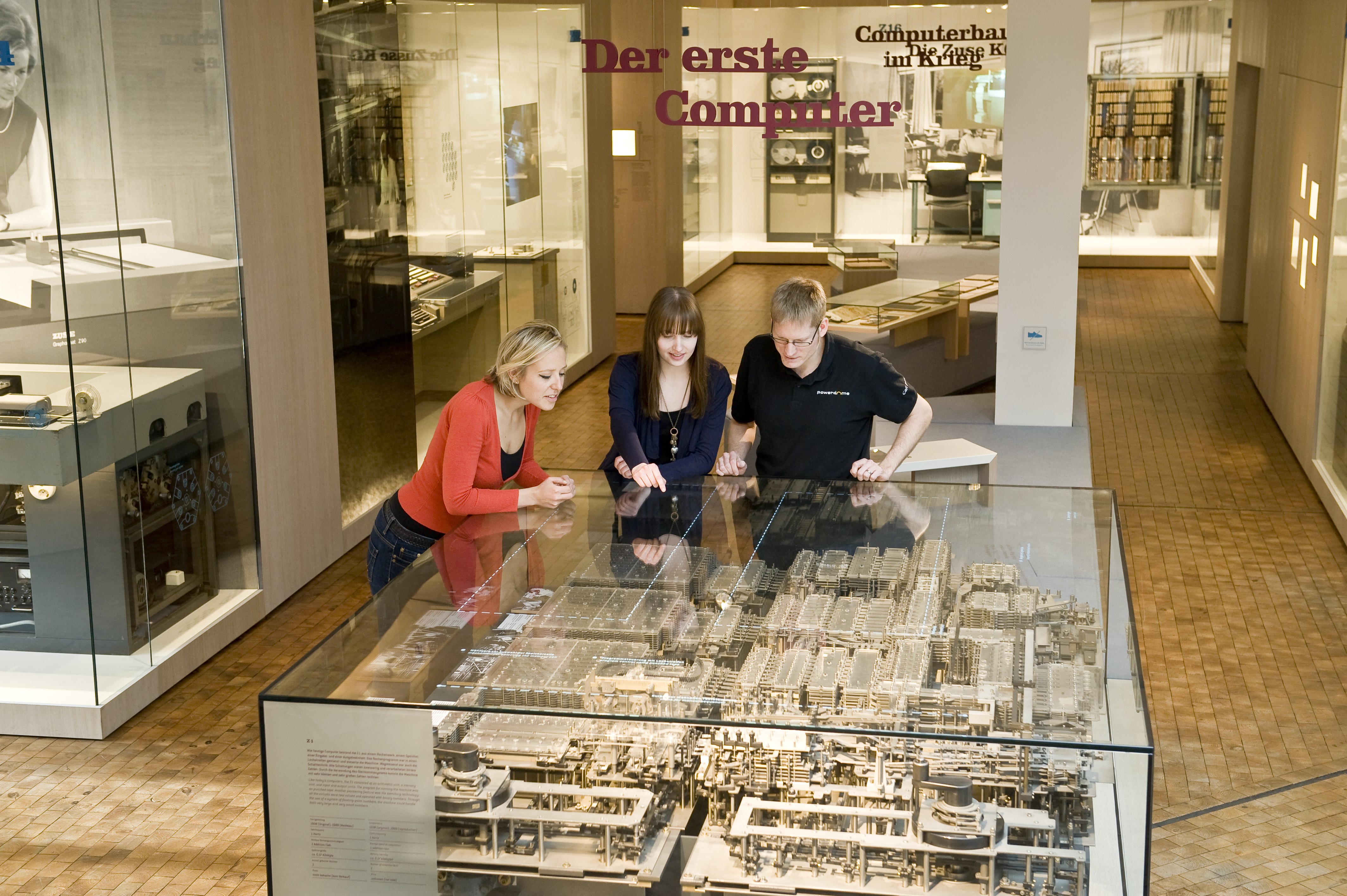 Two women and a man are leaning over a large glass display case containing the Z1's mechanical computing unit.