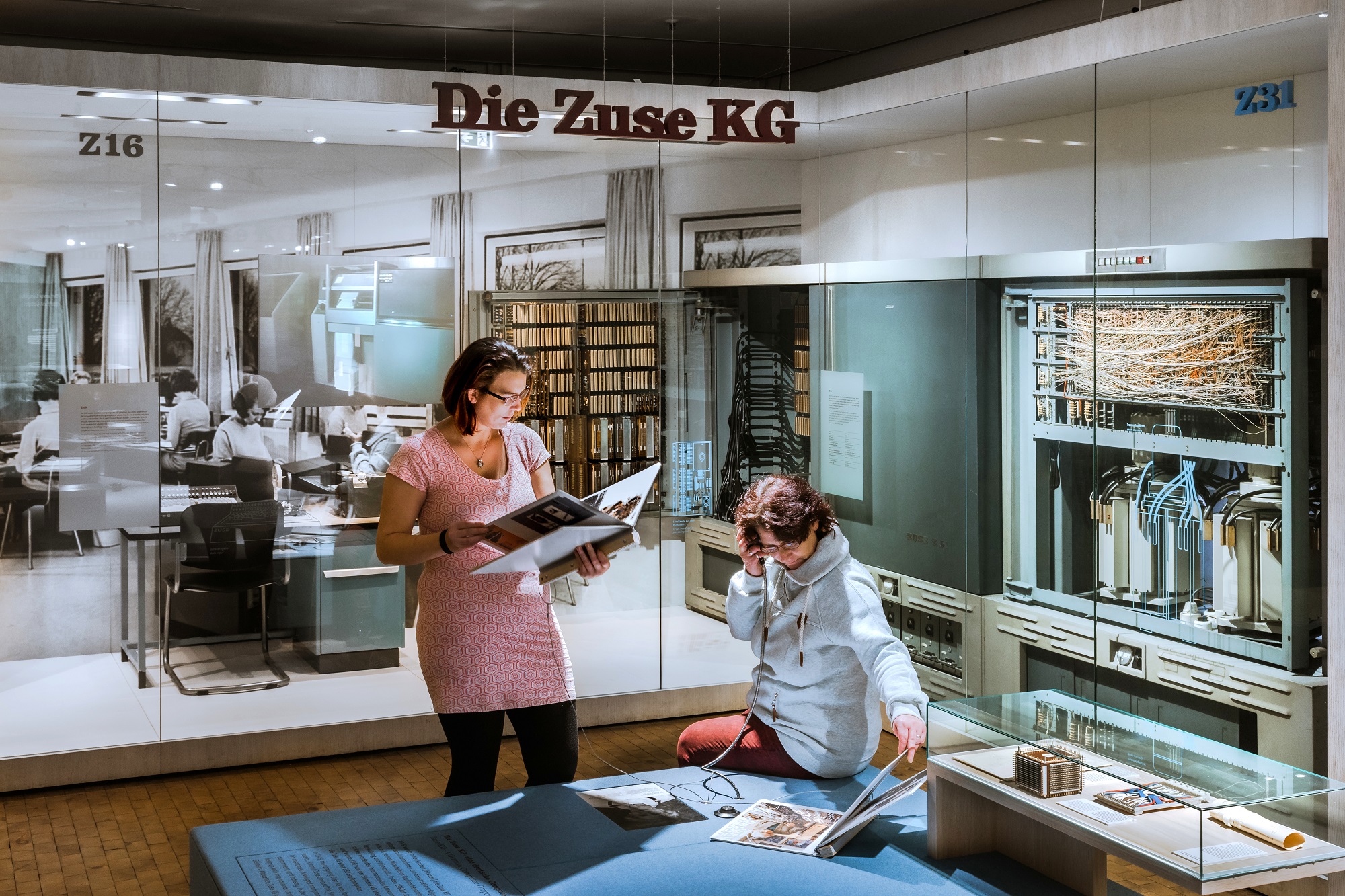 A glimpse into the computer science exhibition: A woman is sitting on a platform, holding the receiver of a listening station to her ear. Another woman is standing next to her, reading from a large notebook. Historic calculating machines are on display in floor-to-ceiling display cases in the background. The words “Die Zuse KG” hang from the ceiling. 