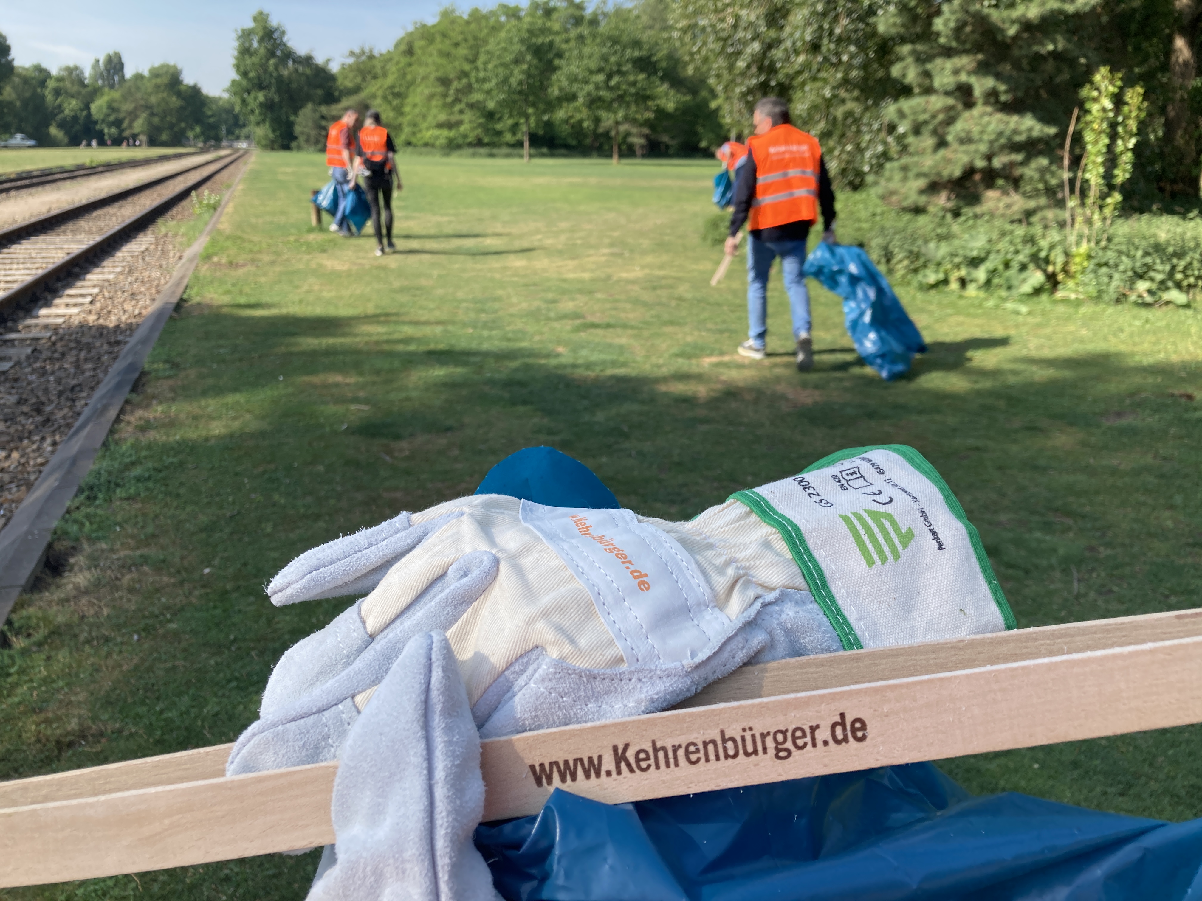 Im Vordergrund Handschuhe und eine Holzzange mt der Aufschrift "www.kehrenbürger.de", im Hintergrund ein Park, in dem Menschen mit orangefarbener Weste und blauen Müllbeuteln umhergehen.