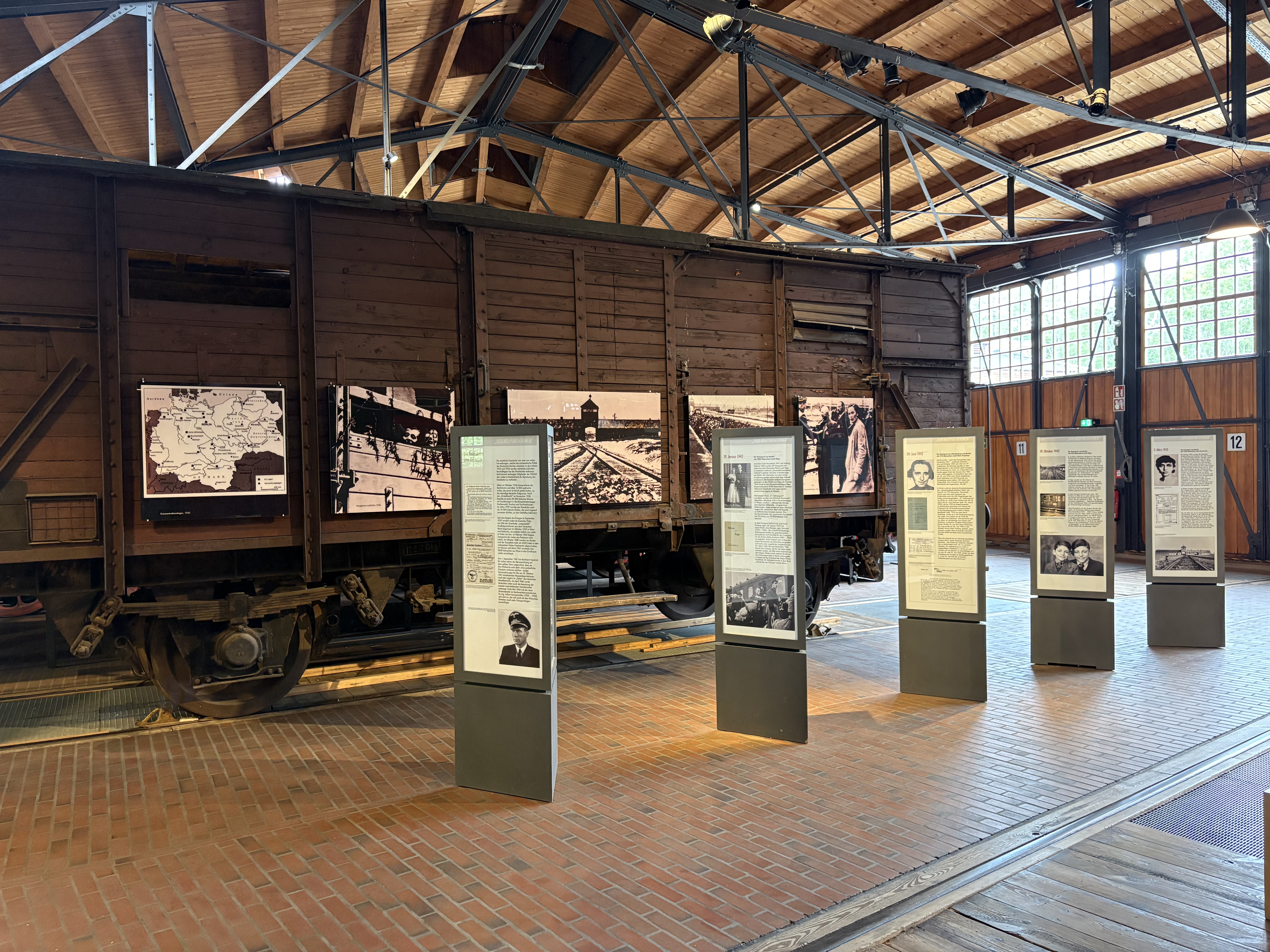 Blick auf einen alten braunen Güterwagen mit Holzaufbau. Auf den Seiten sind Schwarz-Weiß-Aufnahmen aus Konzentrationslagern zu sehen.