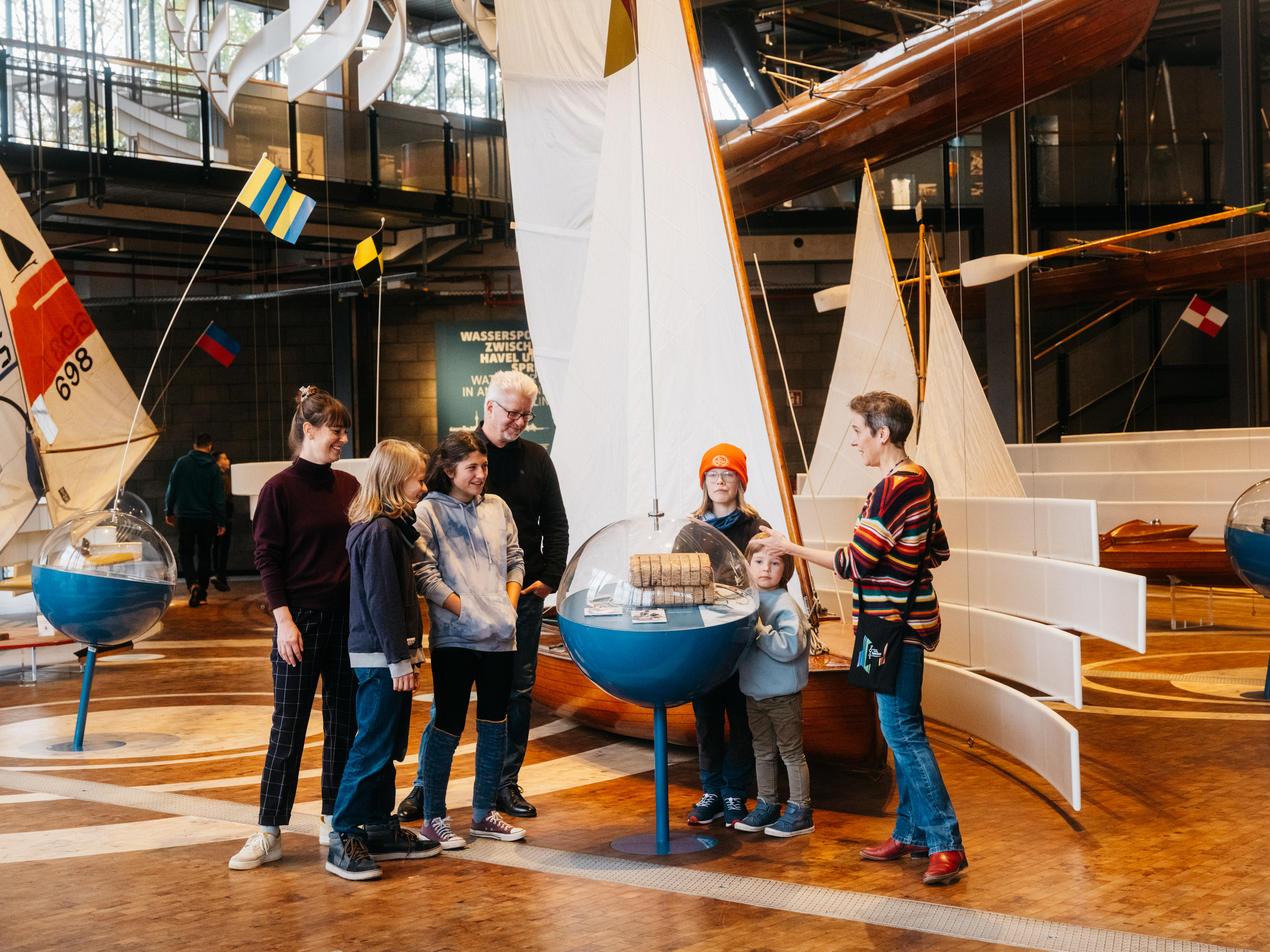 Vier Kinder und drei erwachsene Personen befinden sich in der Schifffahrtsausstellung. Eine Mitarbeiterin des Museum erklärt an einer bojenähnlichen Vitrine ein Objekt. 