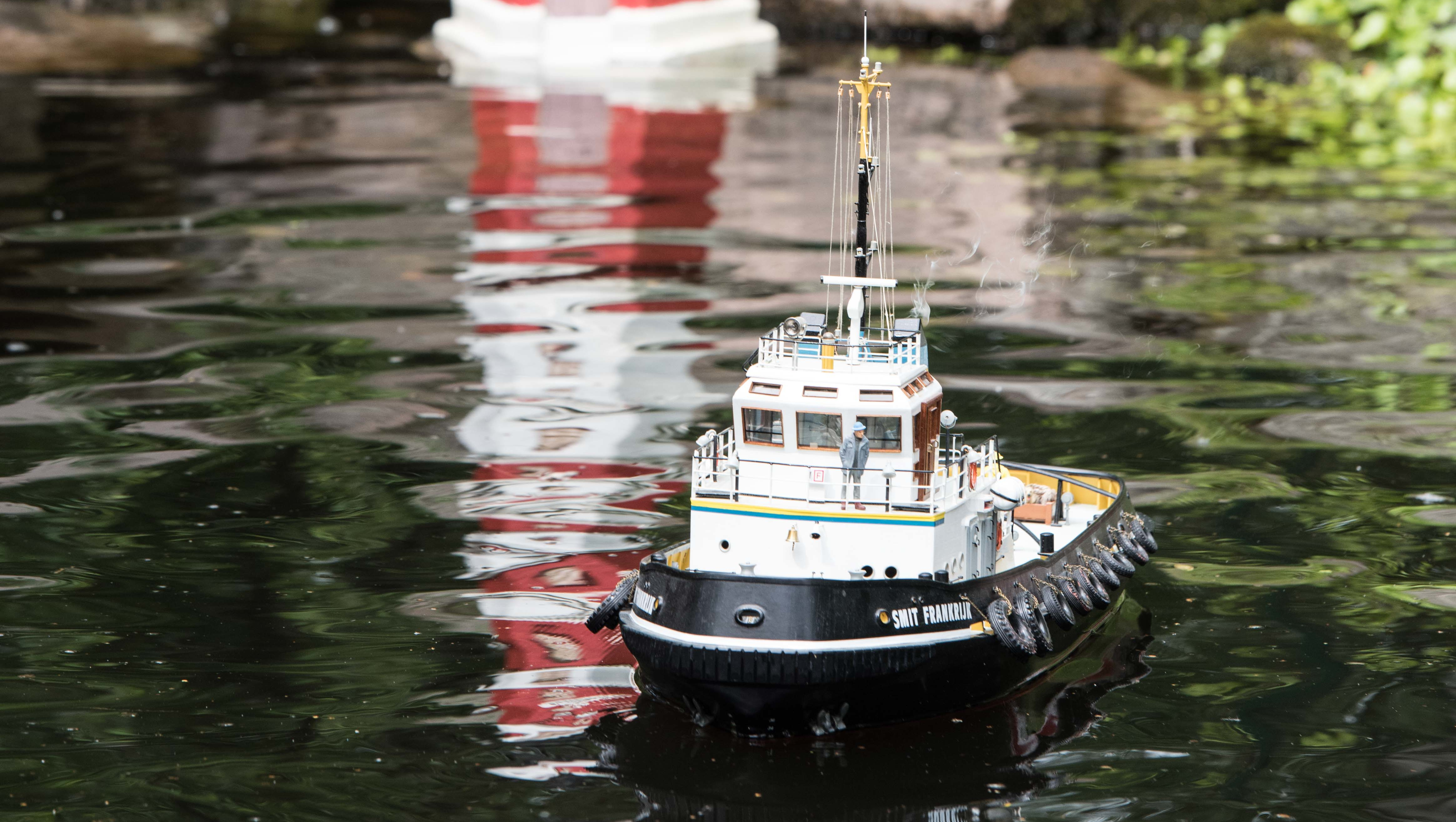 Ein kleines Schiffsmodel fährt auf einem ruhigen Gewässer, im Hintergrund sieht man das Modell eines Leuchtturms, das sich im Wasser spiegelt.
