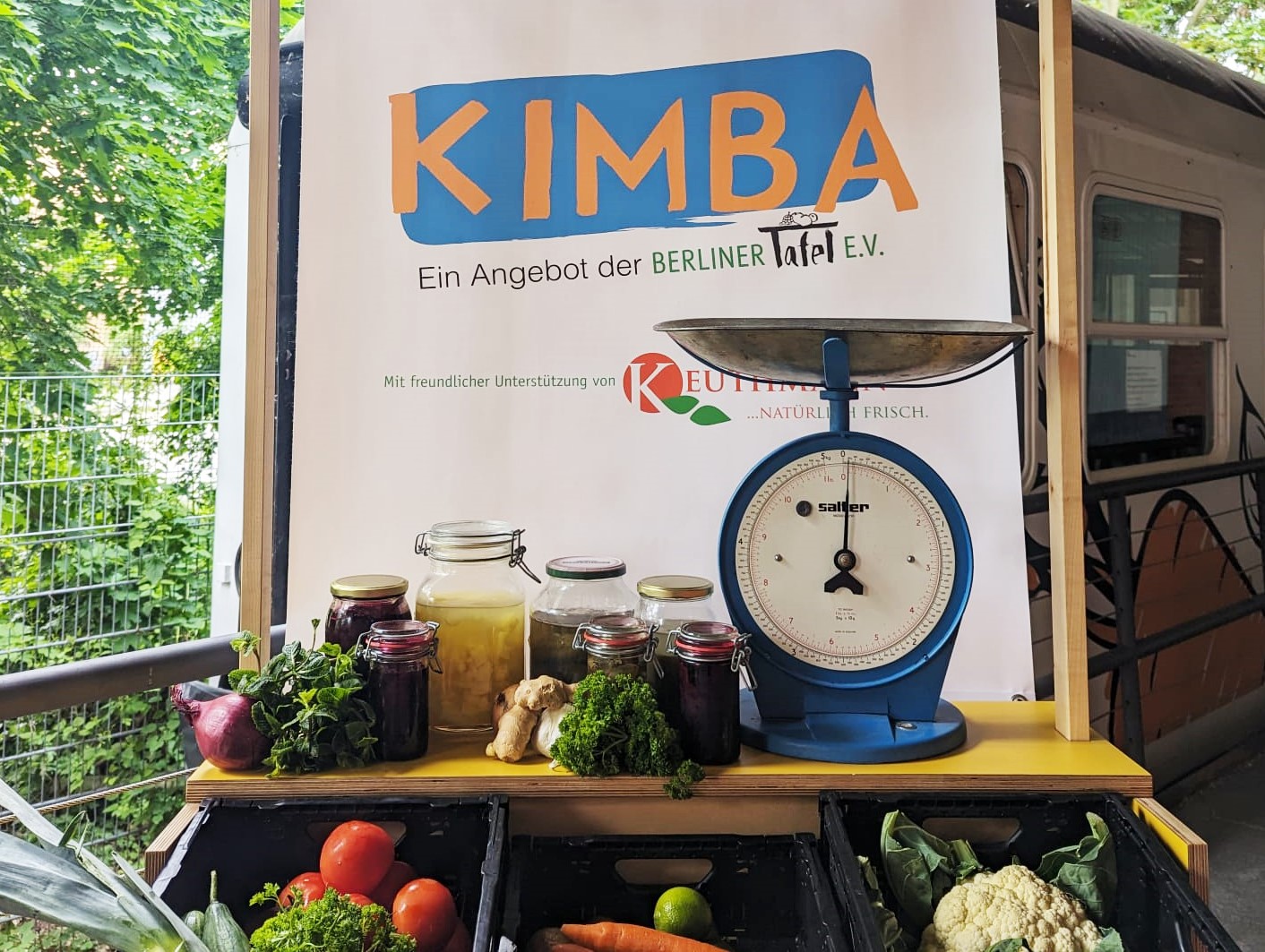 Vor dem KIMBA-Express steht eine Art Markstand mit einer blauen Gemüsewaage und verschiedenen Einweckgläseren mit eingelegtem Gemüse. 