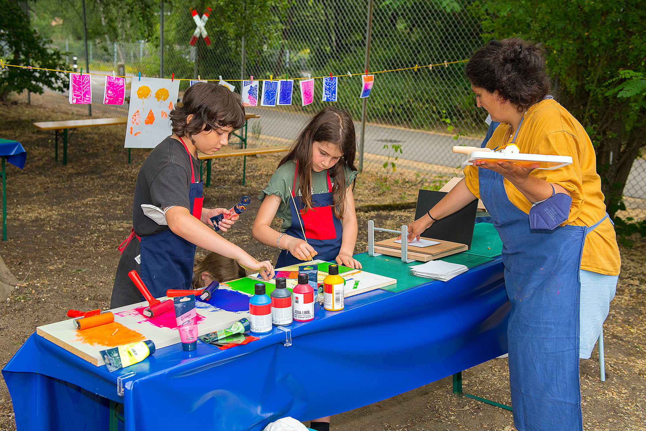 Zwei Kinder und eine Frau stehen an einem Tisch und drucken mit bunten Farben Pflanzenmotive. Im Hintergrund hängen fertige Drucke an einer Leine zum Trocknen.