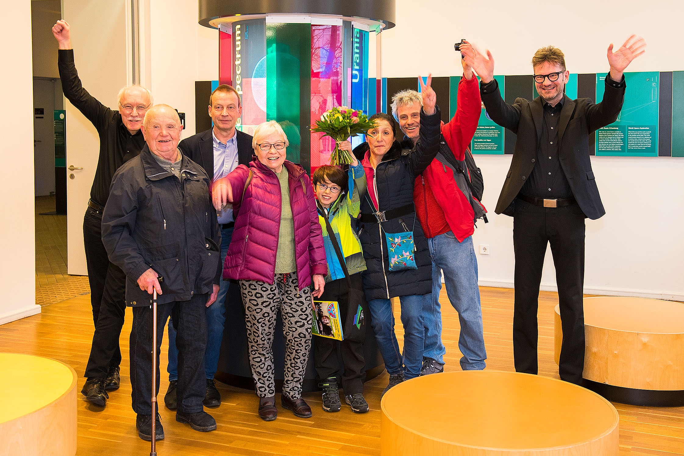 Eine Gruppe von Menschen steht in einem Raum mit bunten Streifen und jubelt in die Kamera.