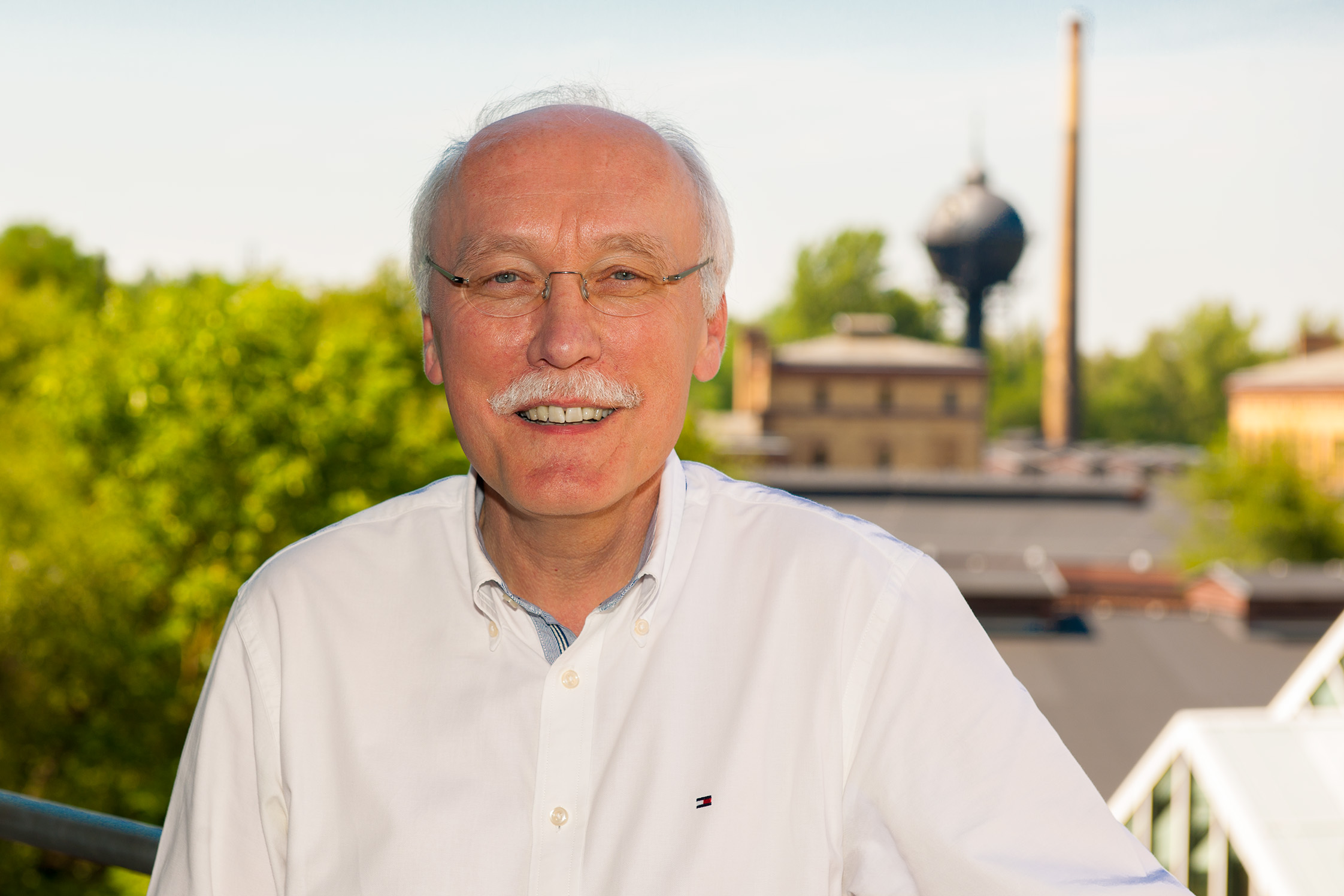 Ein Mann mit weißen Haaren, weißem Schnurrbart und Brille vor dem Museumspark des Deutschen Technikmuseums.