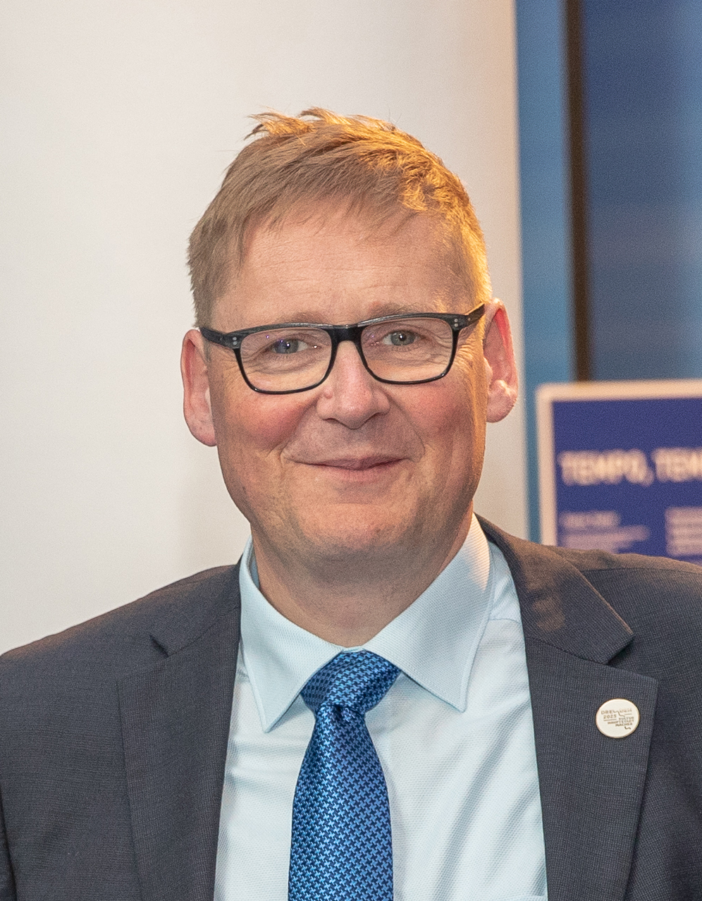 Ein Mann mit blonden Haaren und Brille, Anzug und Krawatte lächelt in die Kamera.