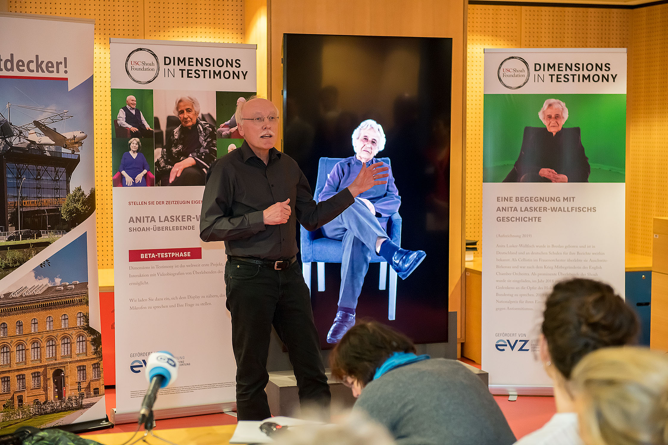 Ein schwarz gekleideter älterer Mannn vor einer Wand mit Banner zu dem Projekt "Dimensions in Testimony"