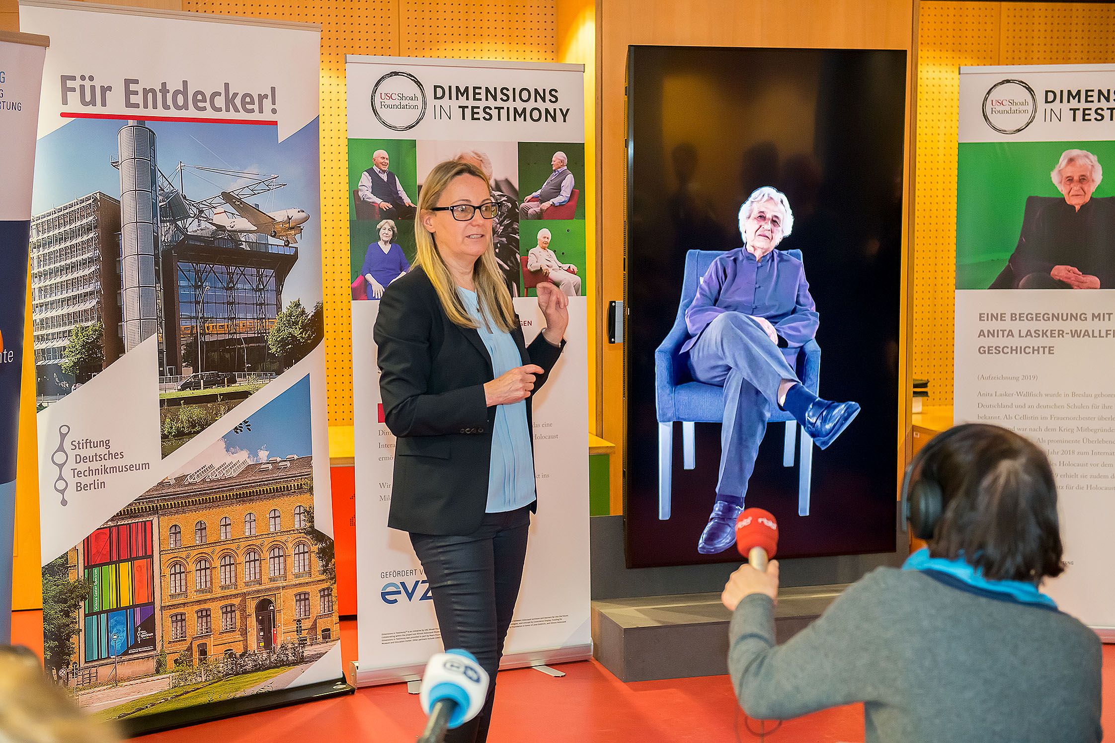 Eine Frau im schwarzen Blazer vor einer Wand mit Bannern des Projekts "Dimensions in Testimony"