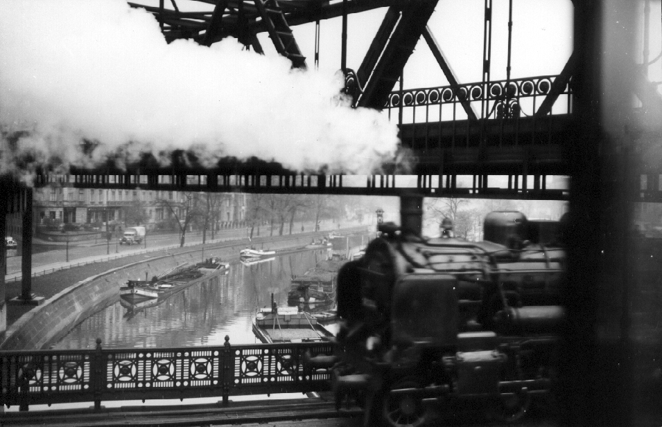 Schwarzweiß-Foto ener Lok, die über eine Brücke fährt, darunter ein Kanal mit Schleppkähnen
