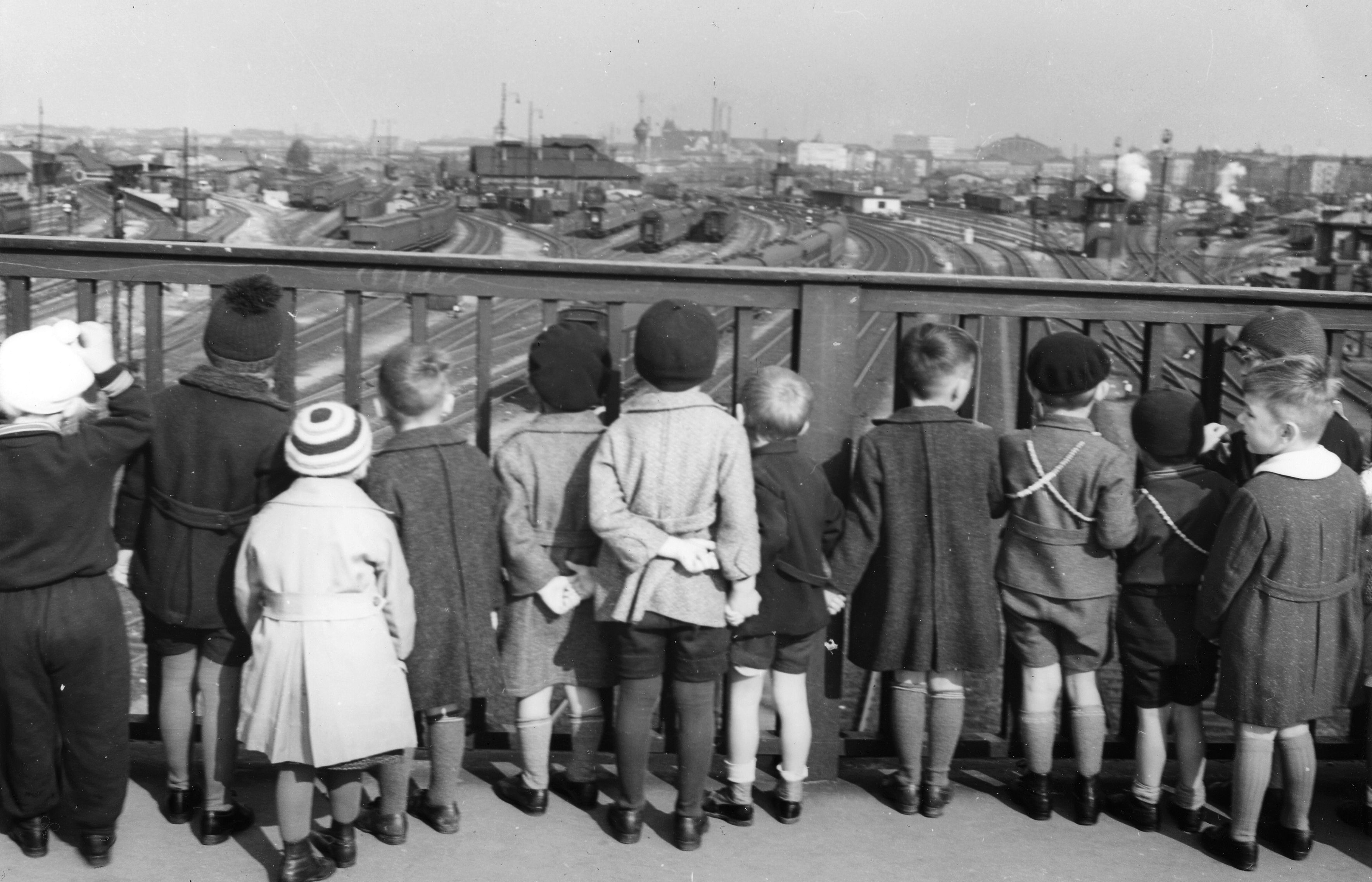 Schwarzweiß-Foto von mehreren Kindern, die mit dem Rücken zum Betrachten auf einer Brücke stehen und ein Gelände mit vielen Schienen anschauen.