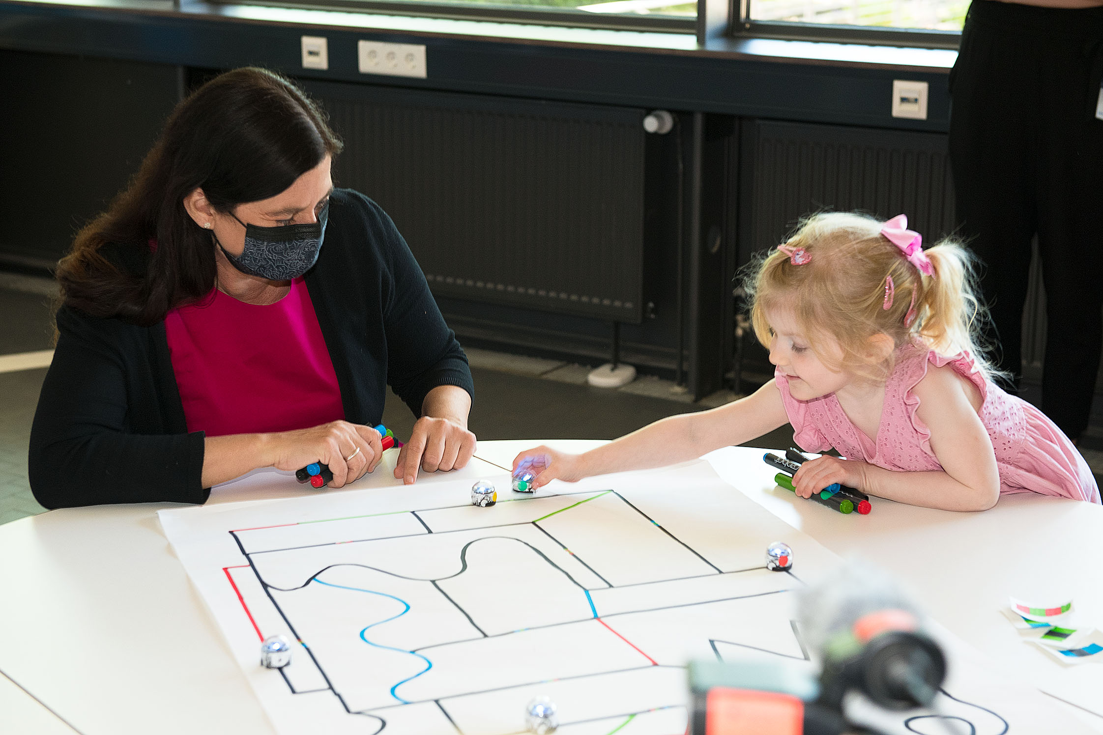 Man sieht eine Frau und ein kleines Mädchen, die an einem Tisch einen kleinen Roboter auf einem Blatt Papier, auf dem eine Linie aufgezeichnet ist, entlangfahren lassen.