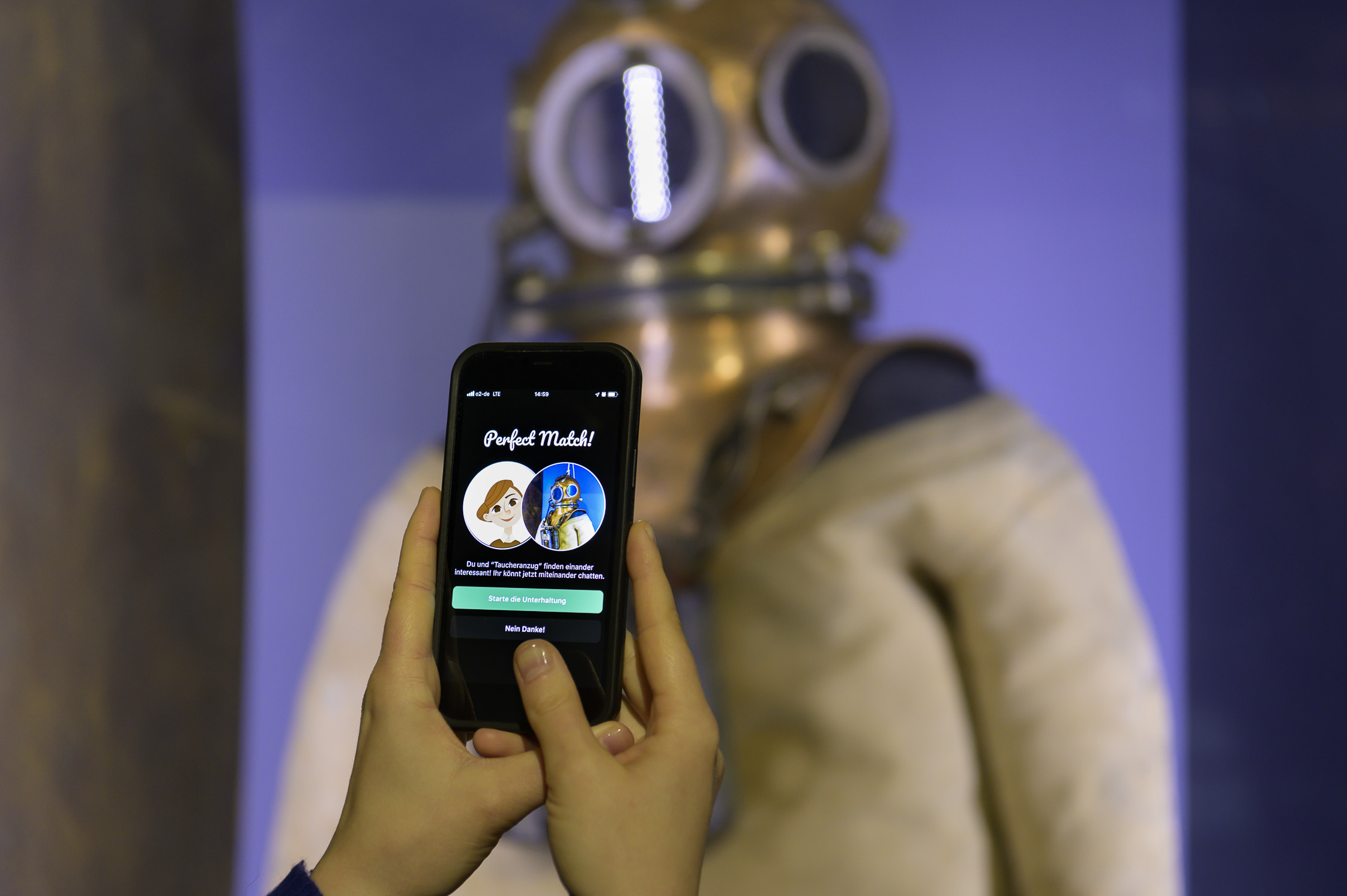 Hände halten ein Smartphone, auf dem Screen steht "Perfect Match", im Hintergrund ist ein Taucheranzug zu erkennen.