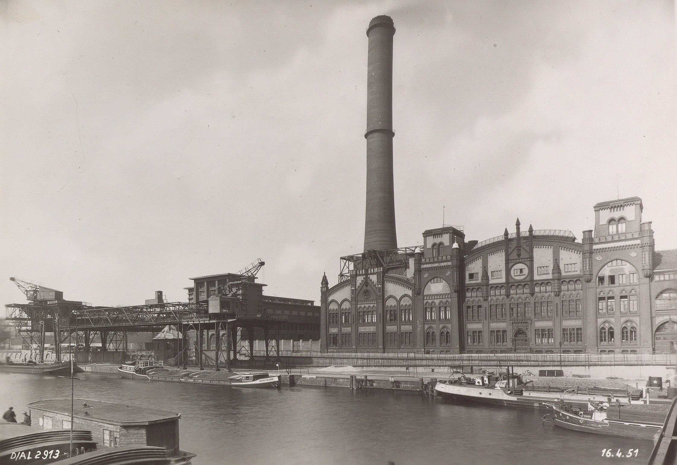 An der Spree befindet sich das Elektrizitätswerk Charlottenburg mit geschwungenen Formen und Rundbogenfenstern. In der Bildmitte ist ein großer Schornstein zu sehen. Die Krananlage für die Entladung der Kohleschiffe befindet sich links im Bild.