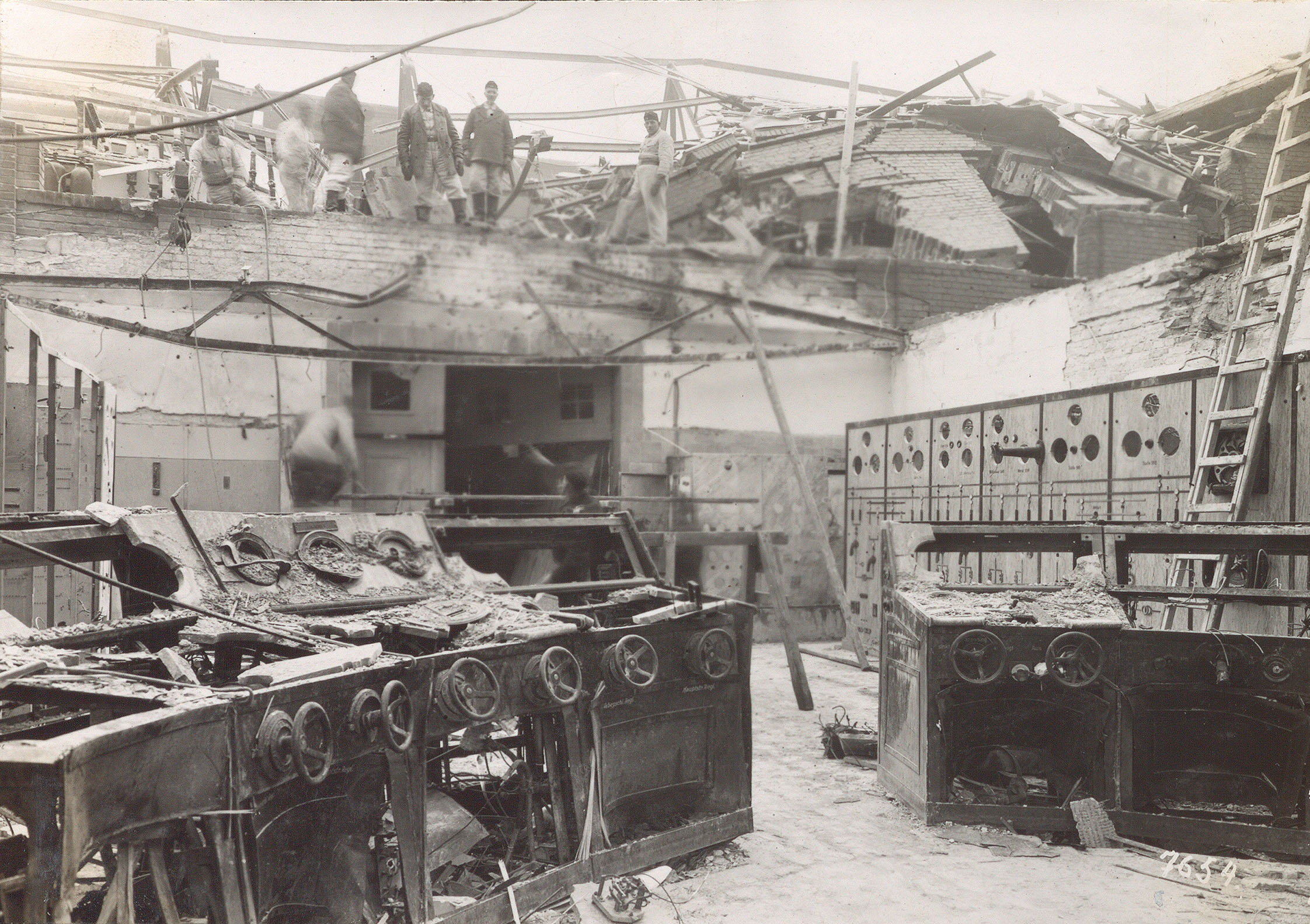 Das Foto zeigt Kriegsschäden am Kraftwerk Spandau 1943/1944. Im Vordergrund des Bildes sind zerstörte Bedienpulte mit Kurbelrädern zu sehen. Die Decke des Raumes ist zerstört. Im darüber liegenden Stockwerk stehen sechs Arbeiter. 