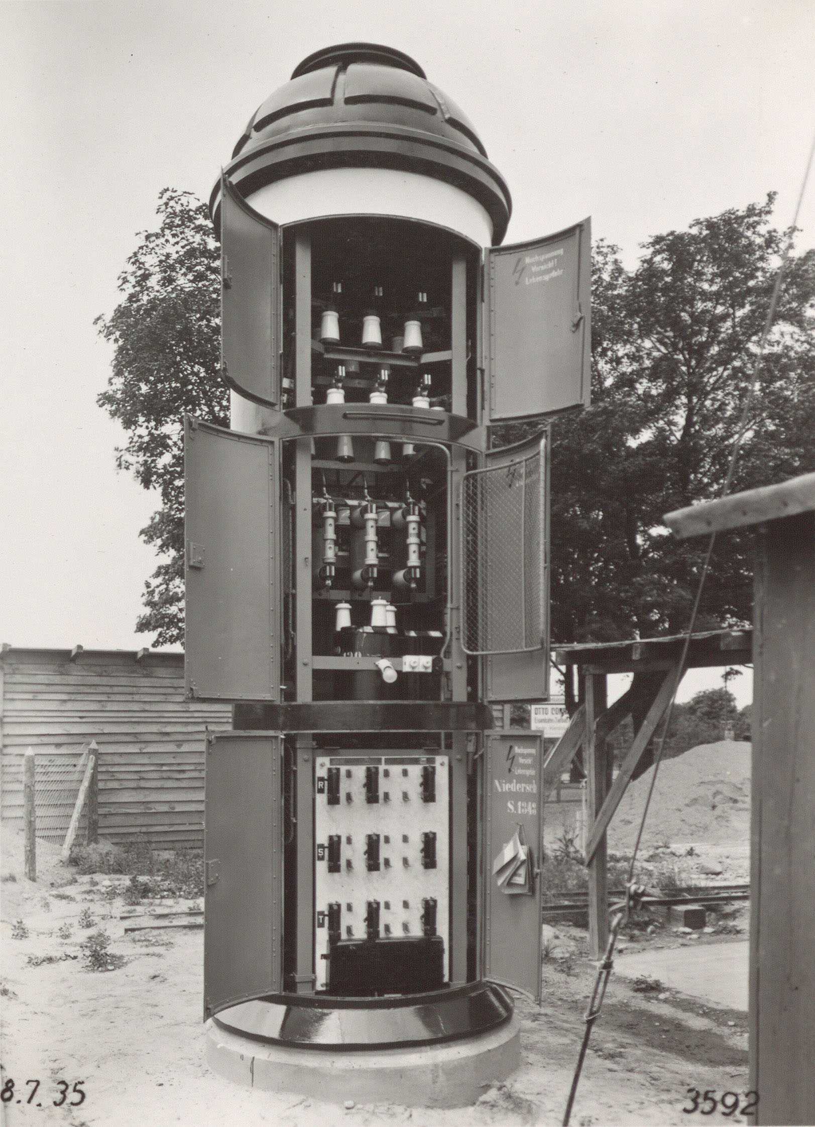 Die kegelförmige Litfaßsäule ist auf einer Seite geöffnet. Auf drei Ebenen befinden sich Türen, hinter denen die Technik des Transformators zugänglich ist. Es ist die Aufschrift „Hochspannung, Vorsicht! Lebensgefahr“ zu lesen.