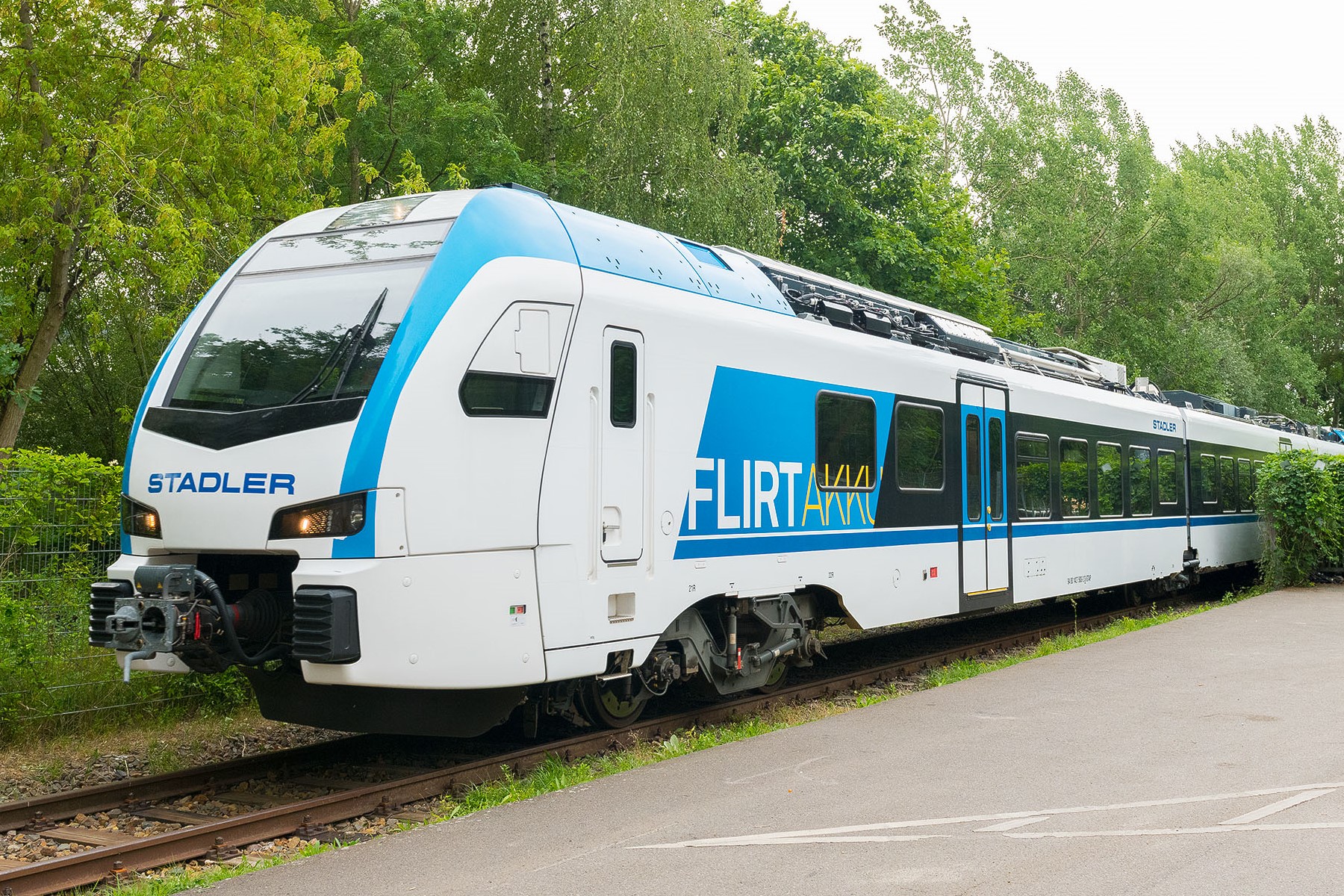 Dreiviertelansicht eines blau-weißen Zugs mit den Aufschriften „Stadler“ und „FLIRT Akku“. Der Zug fährt auf einem Gleis im Grünen, davor ist eine asphaltierte Fläche zu sehen.