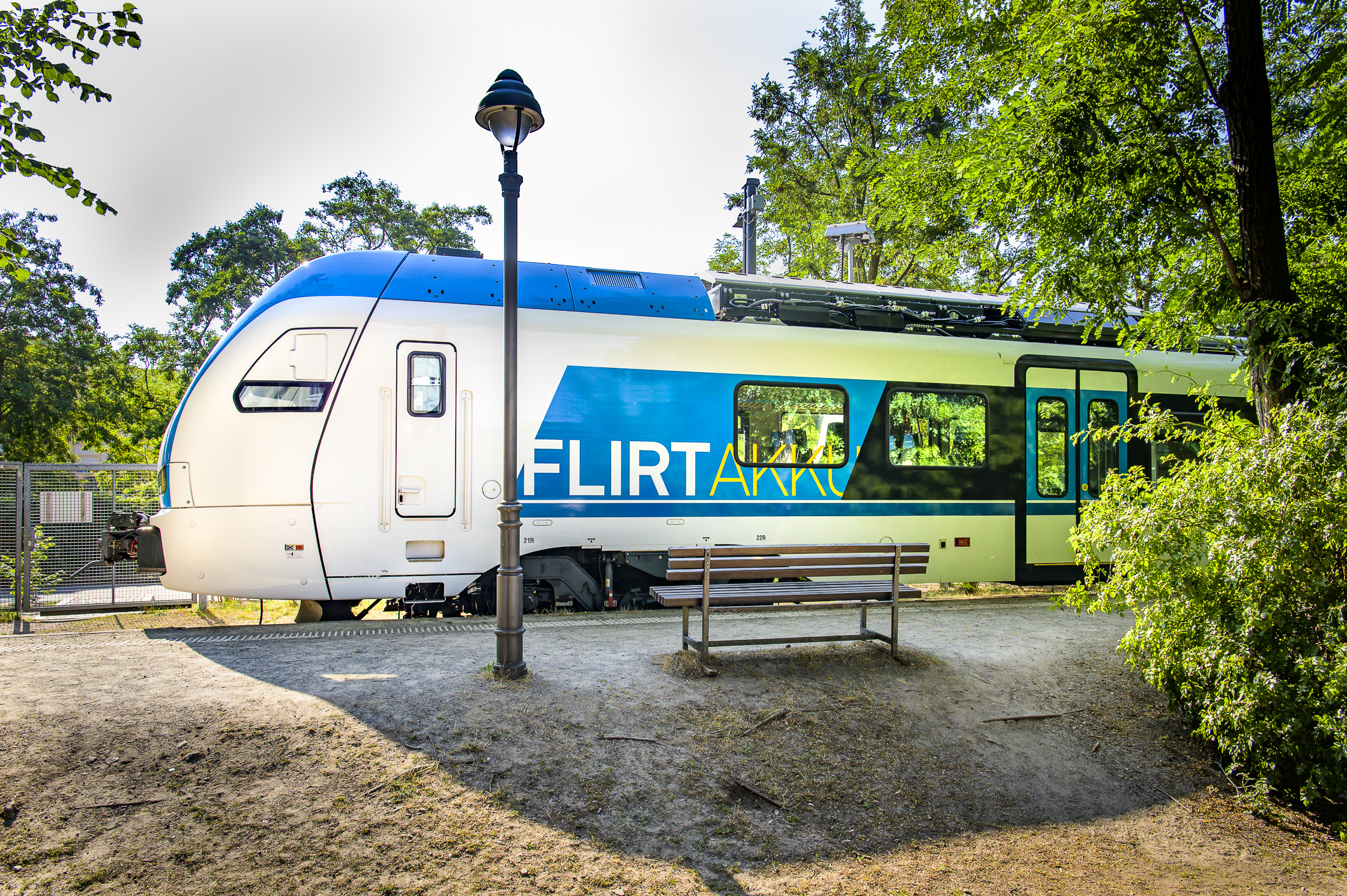 Seitenansicht von einem blau-weißen Zug mit der Aufschrift "FLIRT Akku". Der Zug steht auf einem Bahnsteig im Grünen ein, davor eine Laterne und eine Bank.