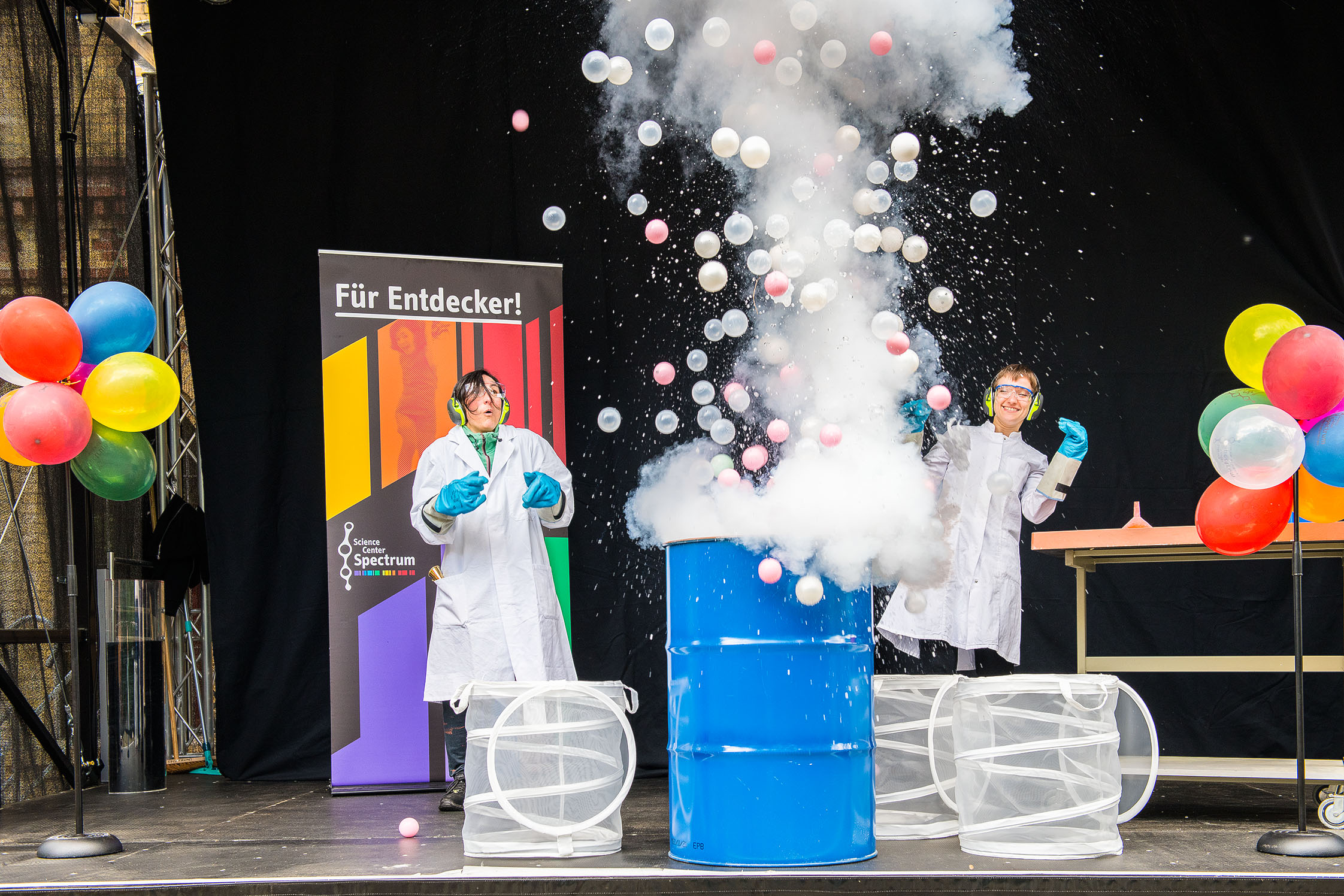 Aus einem blauen Fass explodiert ein weißer Nebel mit bunten Luftballons.