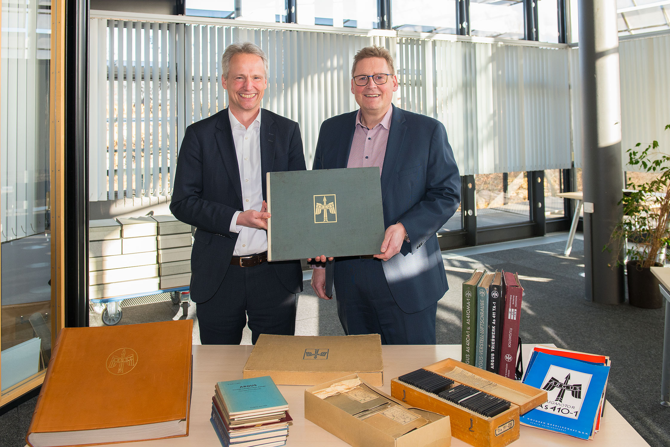 Zwei Männer in Anzügen stehen hinter einem Tisch, auf dem alte Bücher und Unterlagen liegen. Gemeinsam halten Sie ein Buch, auf dessen Vorderseite ein Logo aus Flügeln und einem Pfeil und die Aufschrift „Argus“ zu sehen ist. Im Hintergrund ein lichtdurchfluteter Raum mit großen Fenstern und Jalousien. 