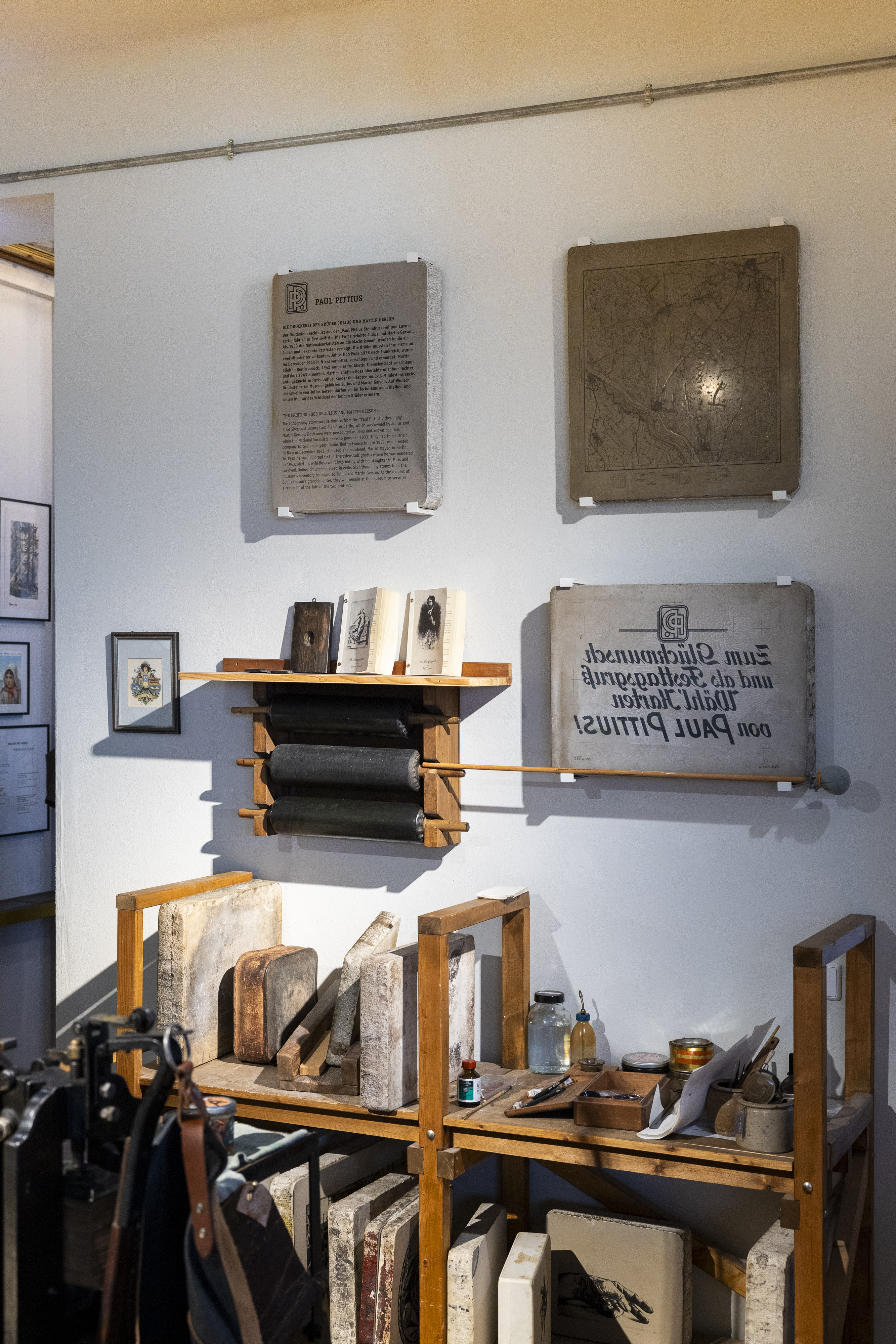 Das Foto zeigt eine Wand, an der mehrere Lithografiesteine hängen, einer davon ist eine Gedenktafel für die Brüder Gerson, Darunter sieht man ein Regal mit verschiedenen Werkzeugen für den Steindruck.