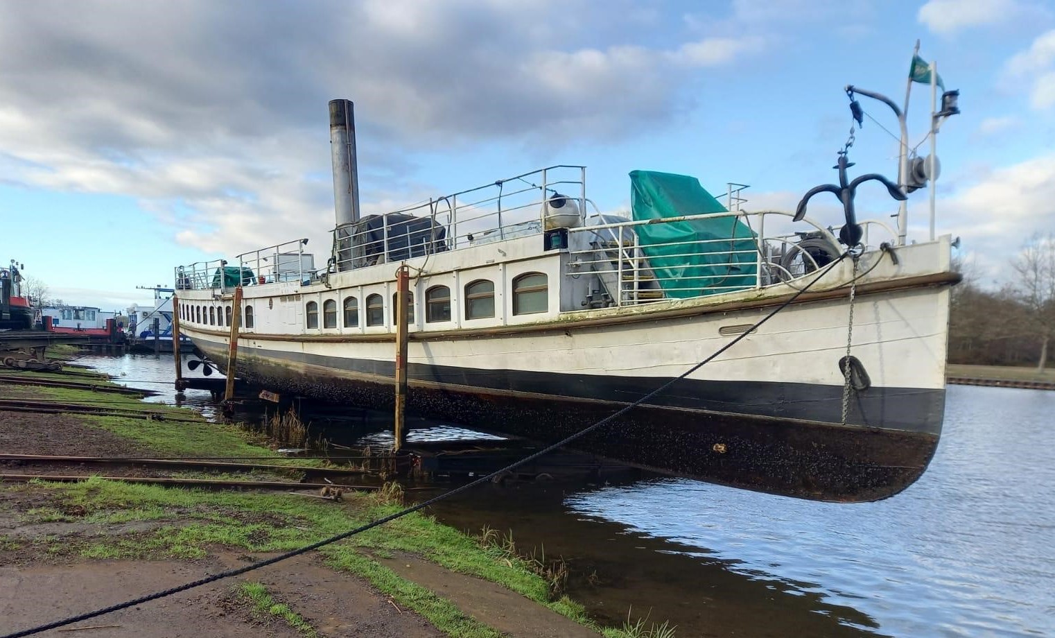 Man sieht ein Schiff am Ufer eines Gewässers, auf einem Gestell vertäut.