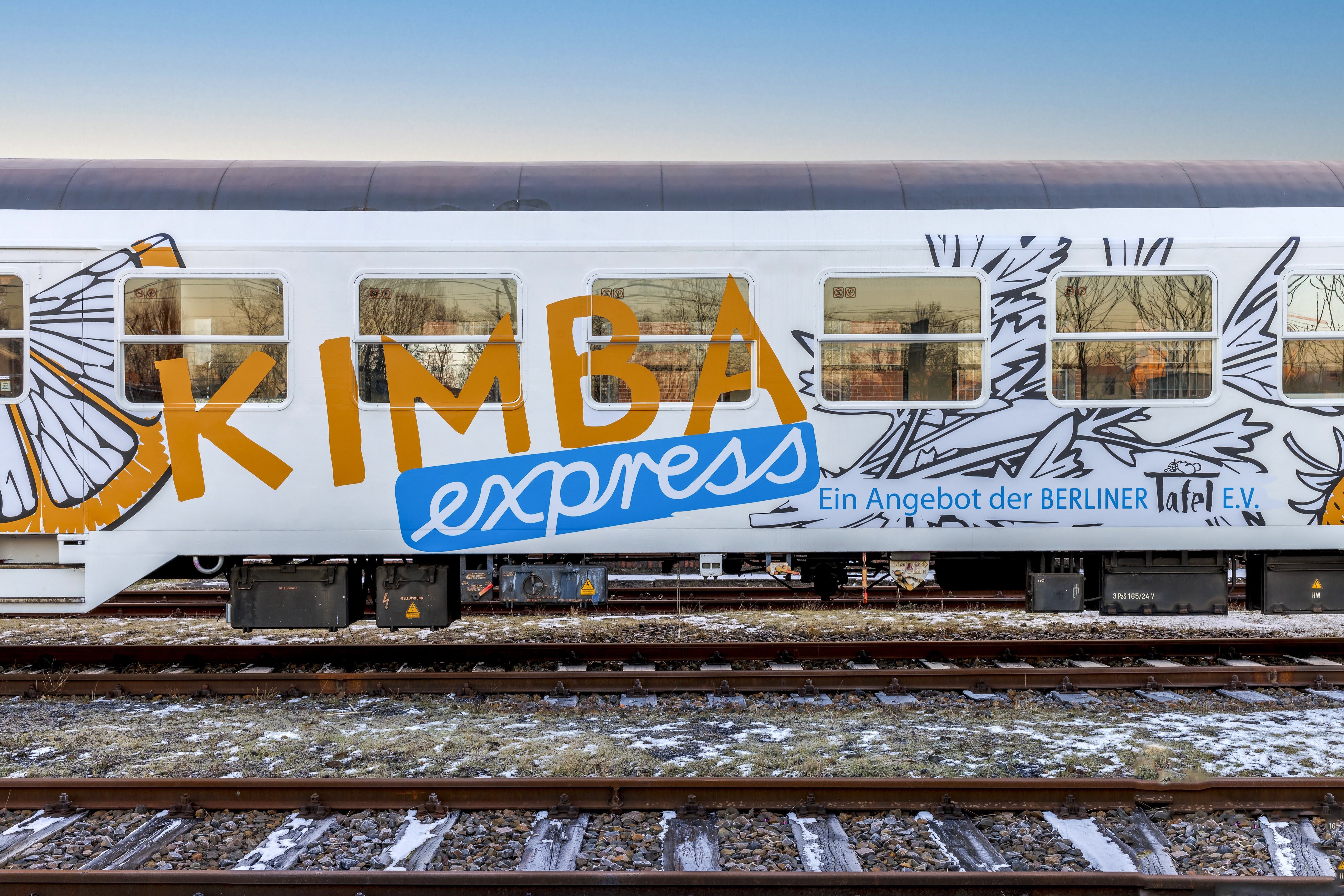 Ein weißer Eisenbahnwagen mit der Aufschrift "KIMBA express" und "Ein Angebot der Berliner Tafel e.V." sowie einer stilisierten Zitronenscheibe und Kräutern.