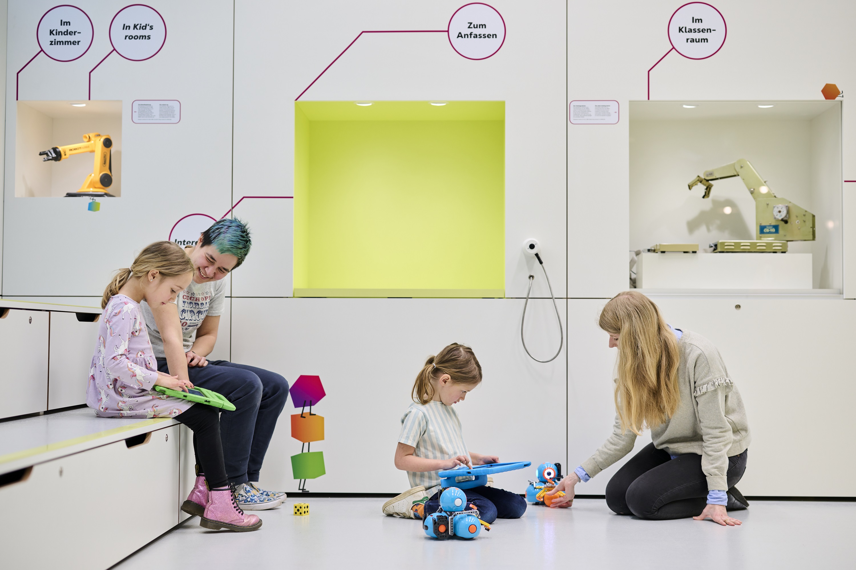 Mehrere Kinder und Erwachsene sitzen auf den Sitzstufen und auf dem Boden eines hell gestrichenen Raums. Sie lesen Bücher und beschäftigen sich mit kleinen Robotern.