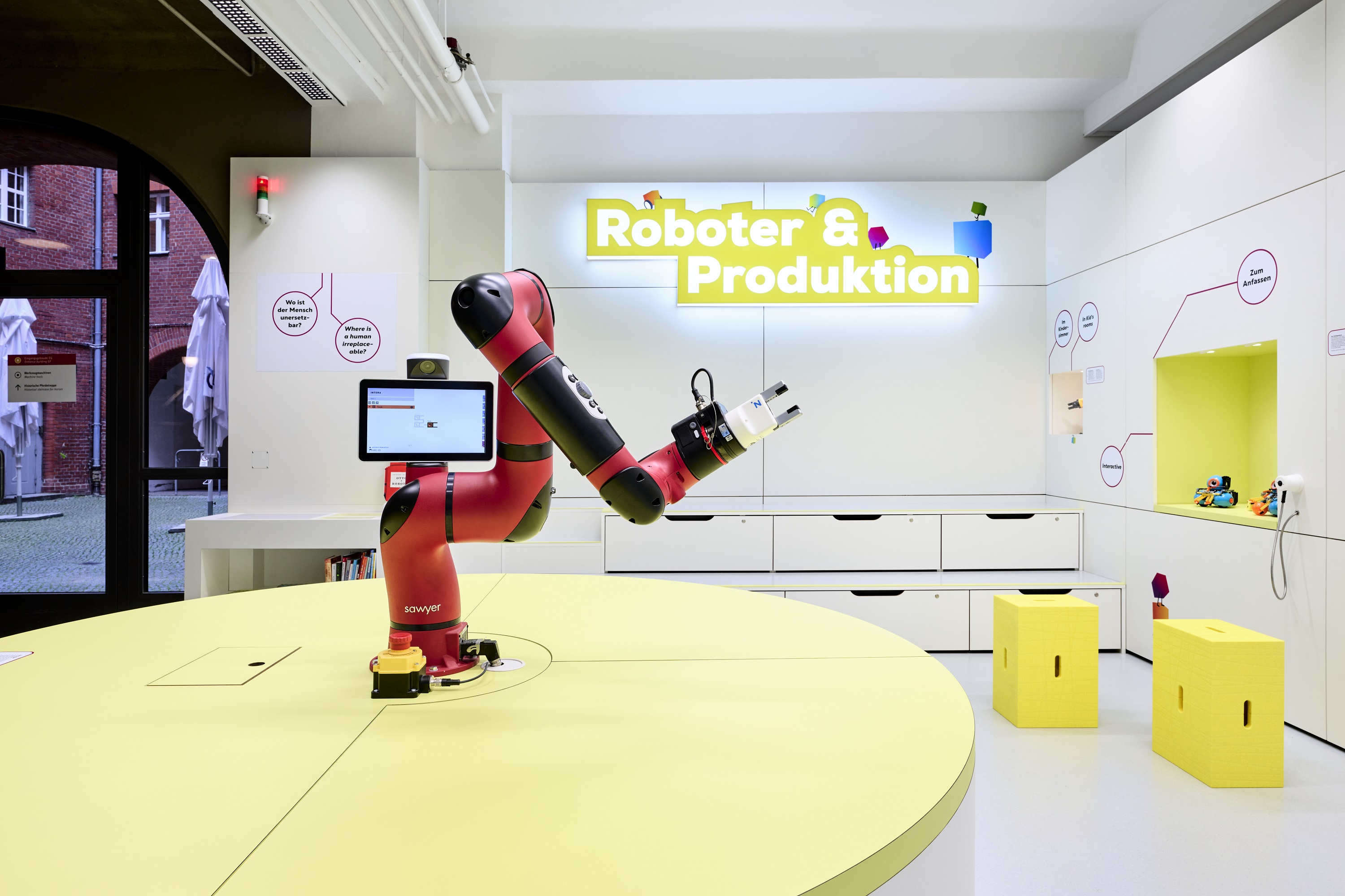 Blick in einen hellgestrichenen Raum in den Farben Weiß und Grün. Auf einem grünen Tisch steht ein rot-schwarzer Roboterarm. Links öffnet sich der Blick in einen Innenhof mit Backsteinmauern.
